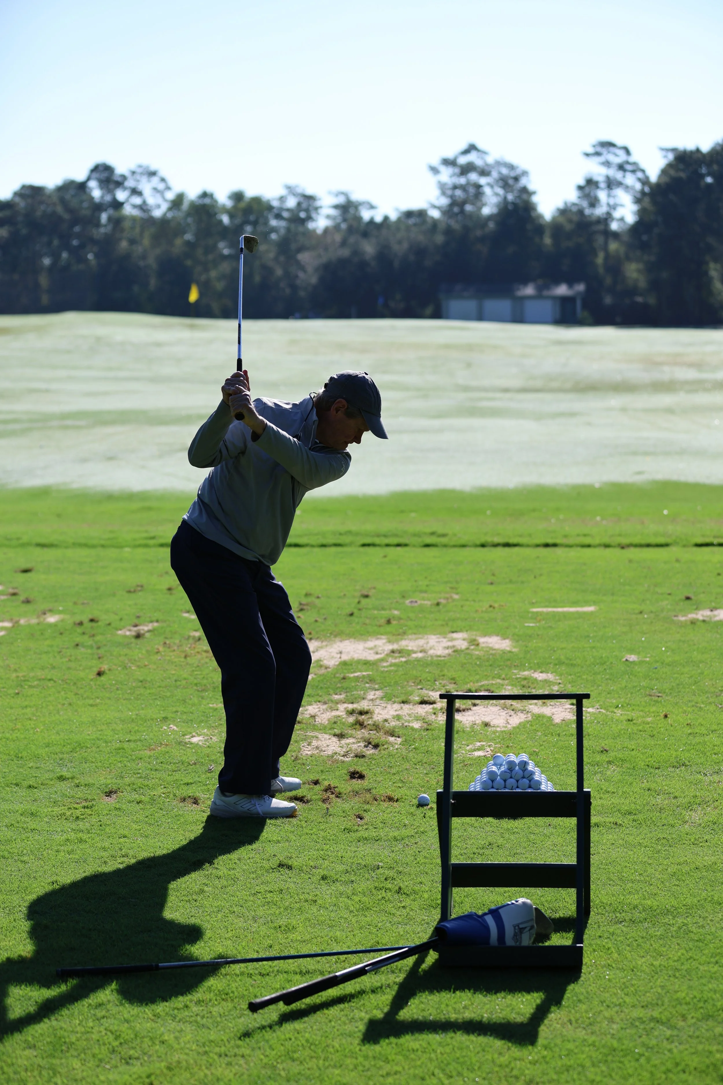 A man practicing golf at a driving range, preparing to swing a golf club with a golf ball on the ground. A basket of golf balls and another golf club are nearby on the grass.