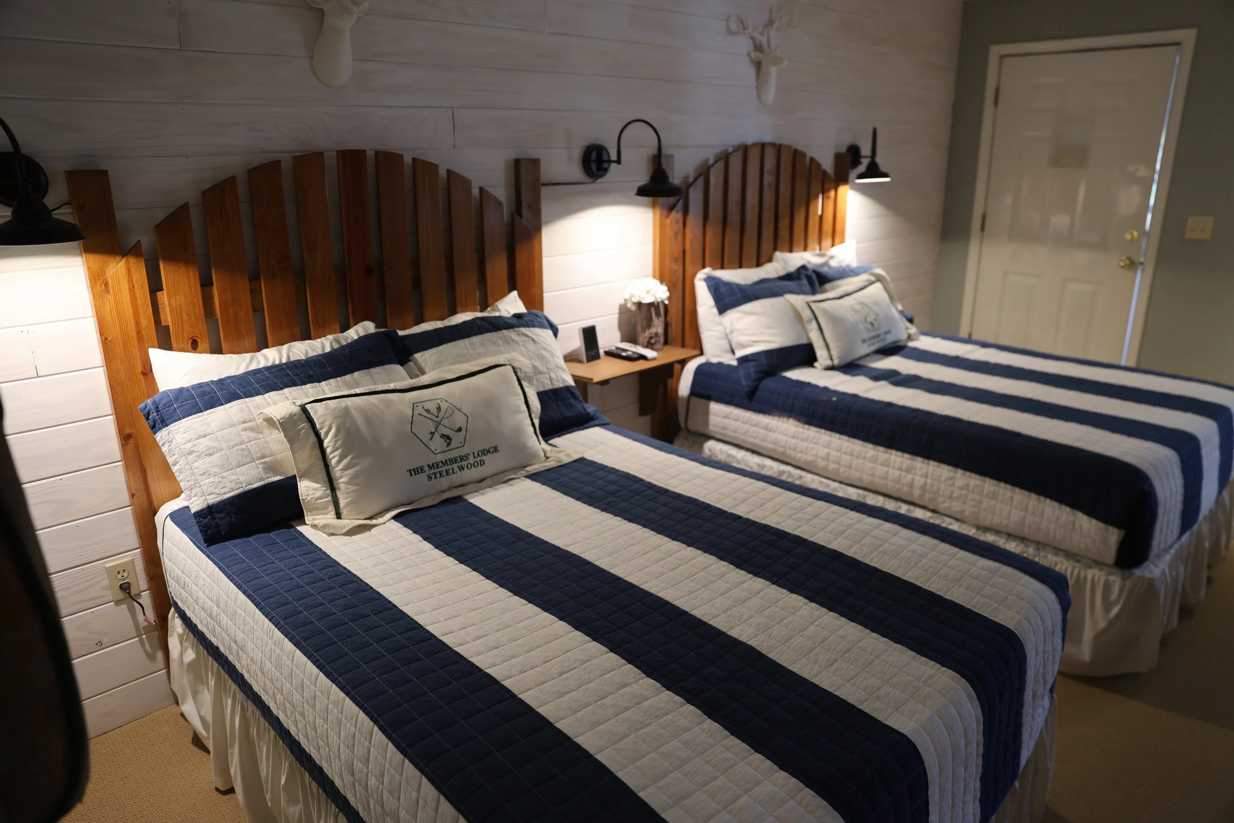 Two beds with matching blue and white striped quilts in a cozy bedroom with wooden headboards and wall-mounted lamps.