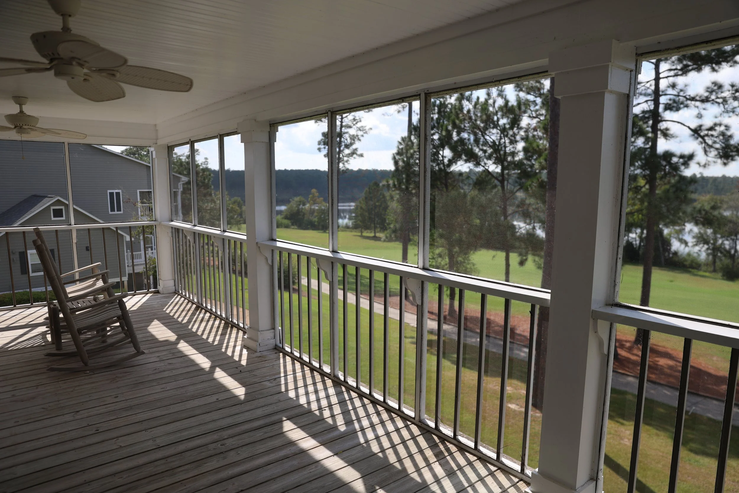 A porch with wooden flooring, two rocking chairs, ceiling fans, white railings, and screens, overlooking a grassy yard with trees and a lake in the distance.