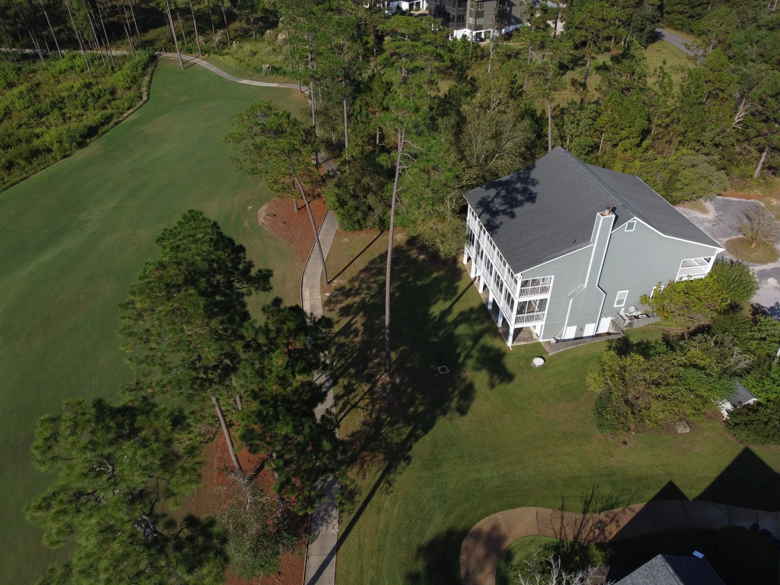 Aerial view of a light gray house with balconies surrounded by green trees, a curved grassy lawn, and a paved walkway.