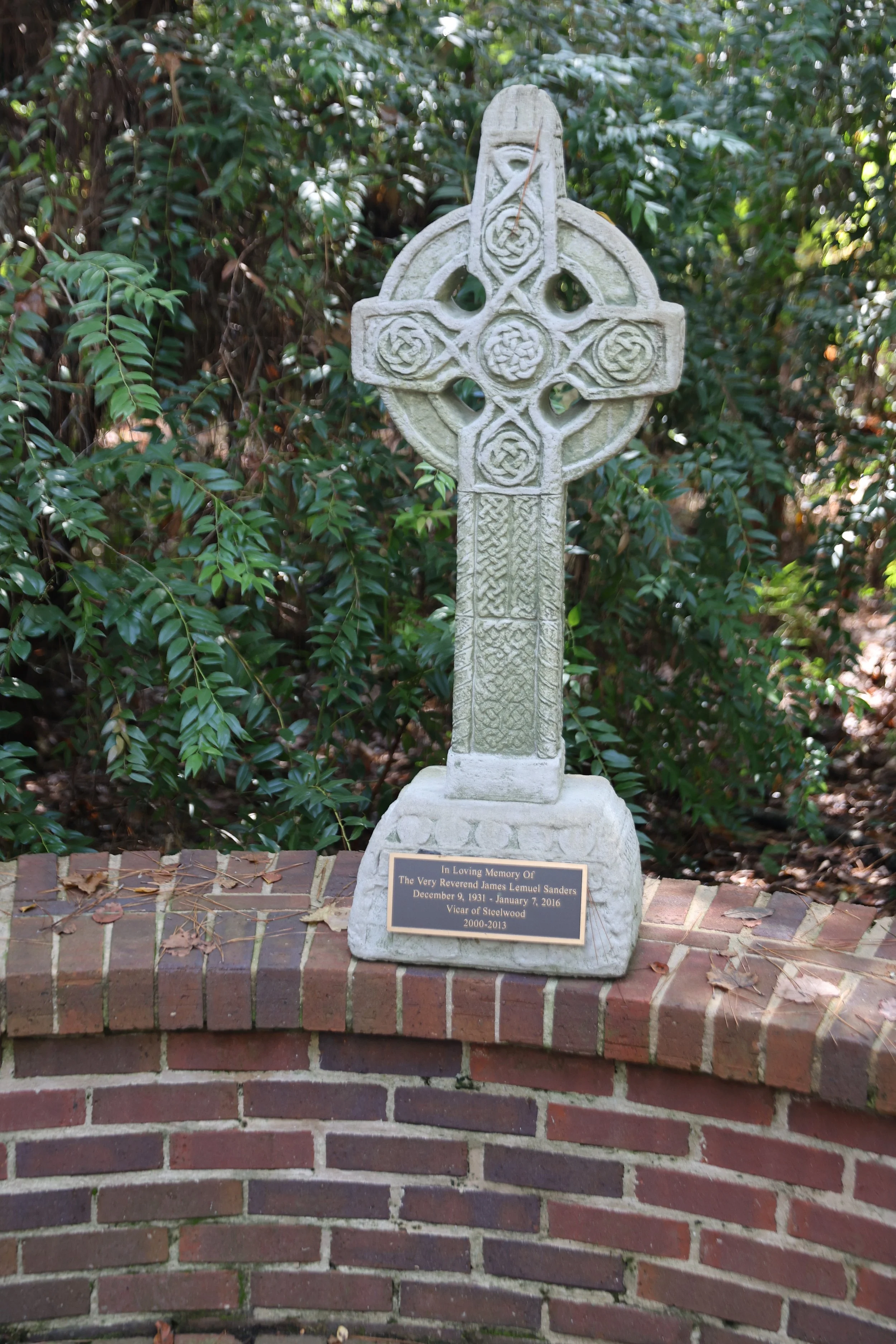 A Celtic cross memorial stone with a plaque at its base, placed outdoors against a background of greenery and bushes. The plaque reads: "In Loving Memory Of The Very Reverend James Lemuel Sanders December 9, 1931 - January 7, 2016 Vicar of Steelwood 