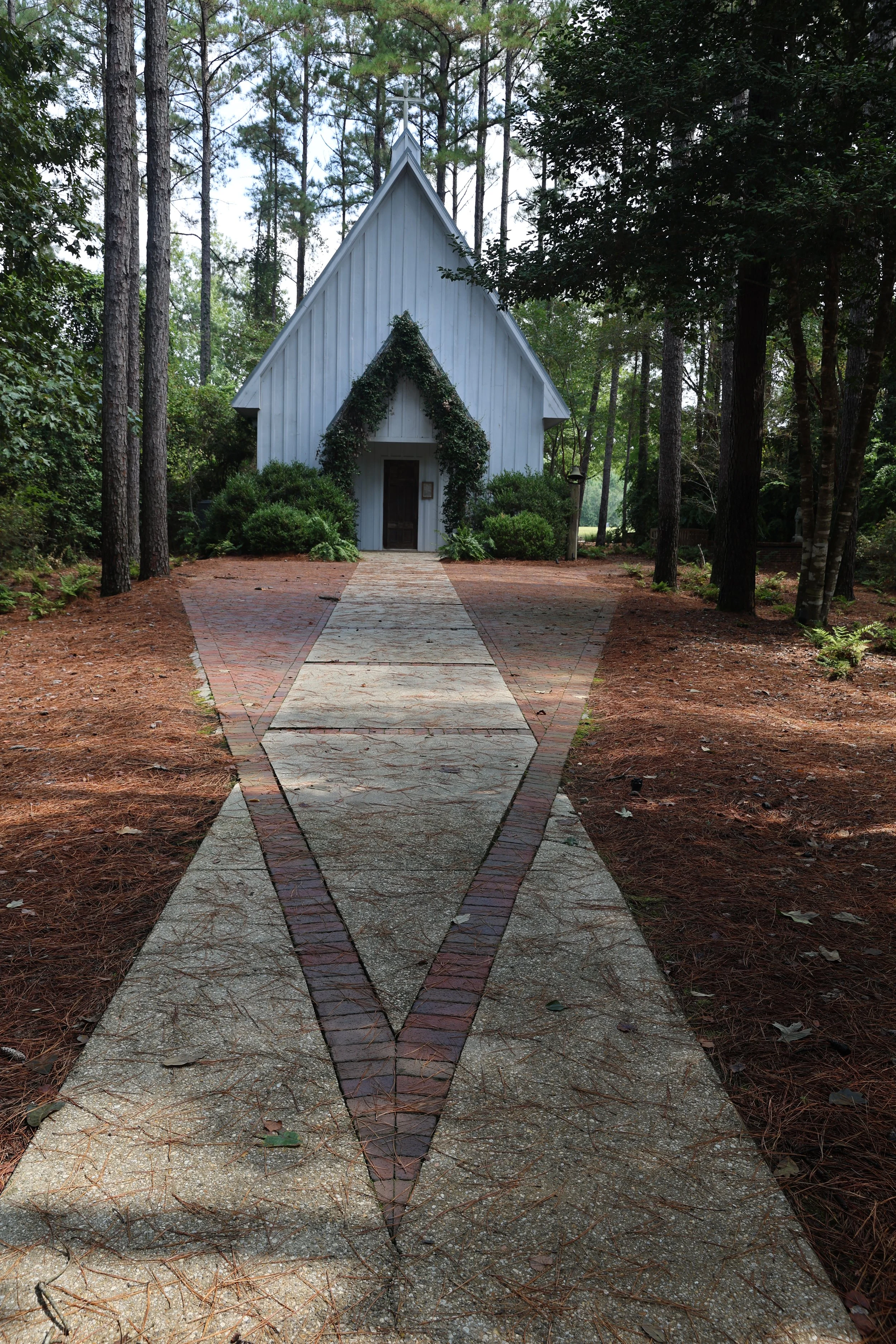 A small white church with a sharp, triangular roof, surrounded by tall trees and bushes, with a paved walkway leading to the entrance.