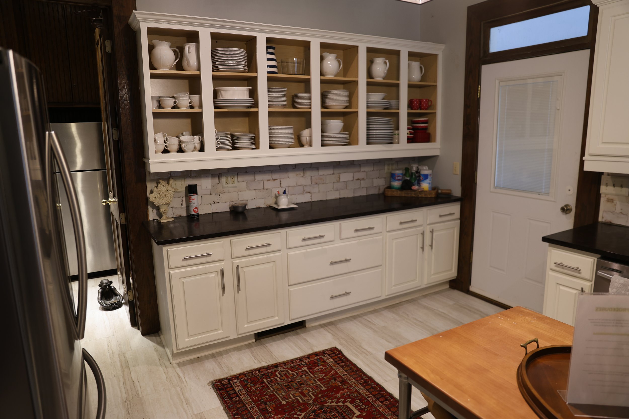 Kitchen with white cabinets and open shelving filled with white dishes, a brick backsplash, and a black countertop. There is a refrigerator on the left and a wooden table on the right. A door with a window is on the right side.