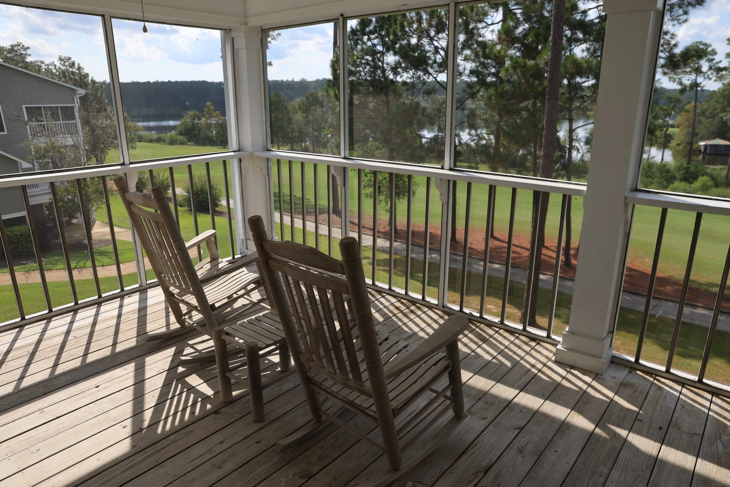 View from a screened porch with two wooden rocking chairs overlooking a grassy landscape with trees, a lake, and a partly cloudy sky.