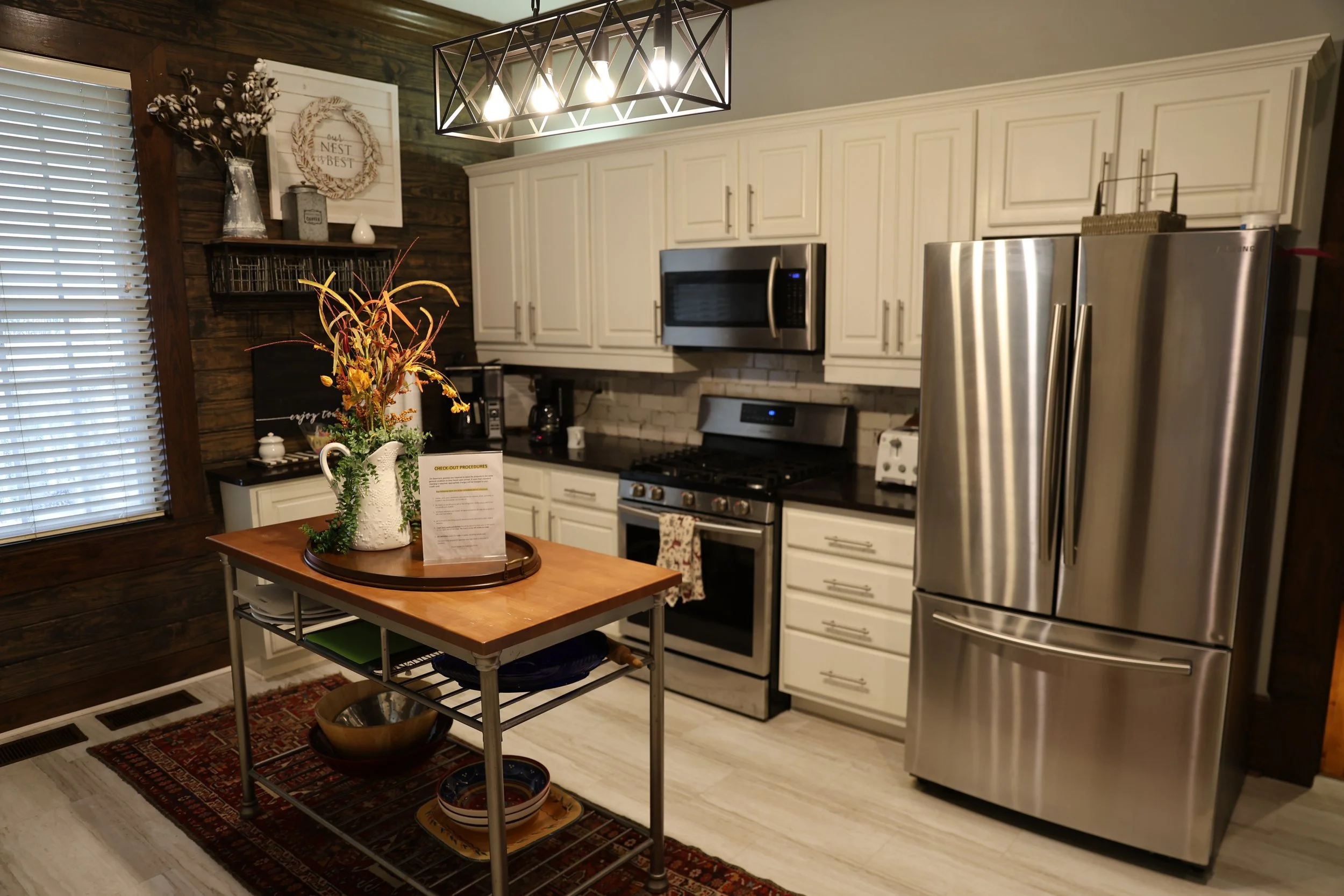 A modern kitchen with white cabinets, a stainless steel refrigerator, a black countertop, and a small island table with a flower arrangement and a menu card, all under a geometric light fixture.