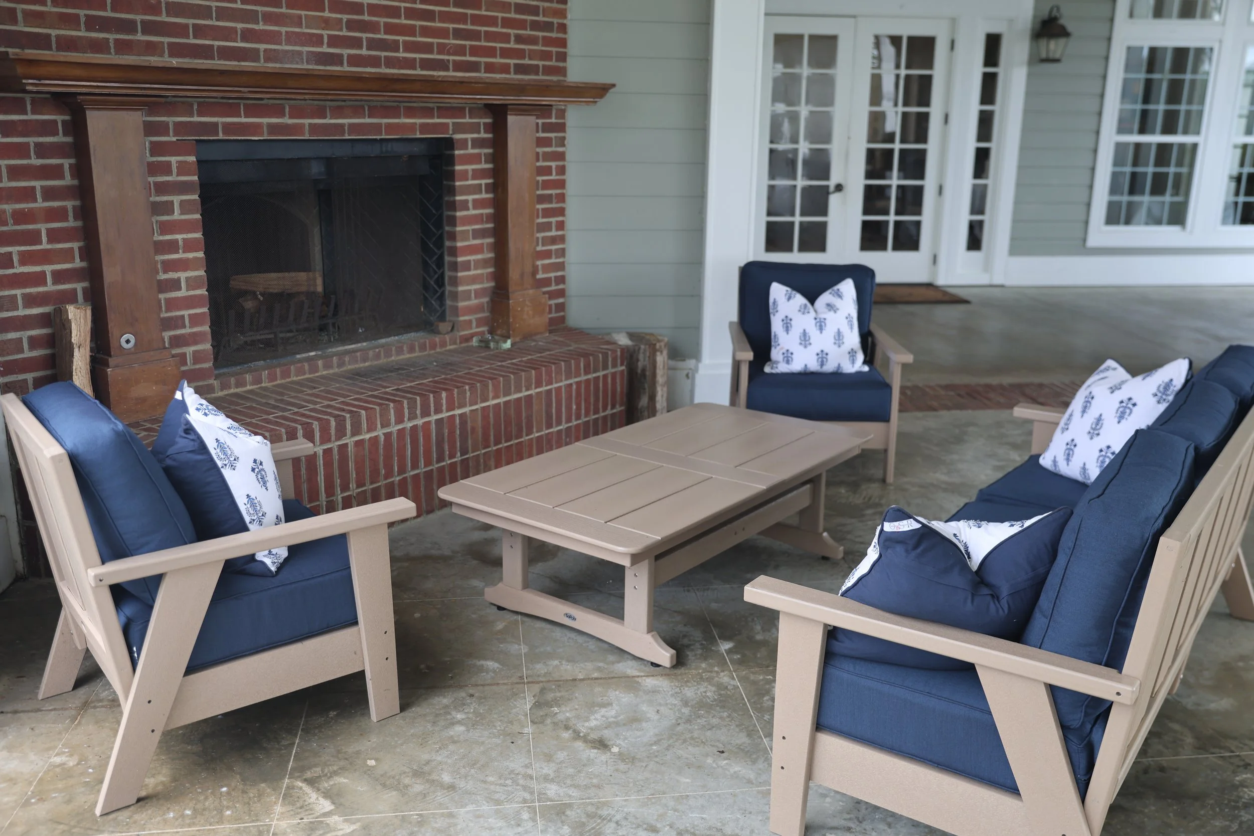 Outdoor patio area with blue cushioned chairs and pillows around a beige wooden table, brick fireplace, and sliding glass door leading into a house.