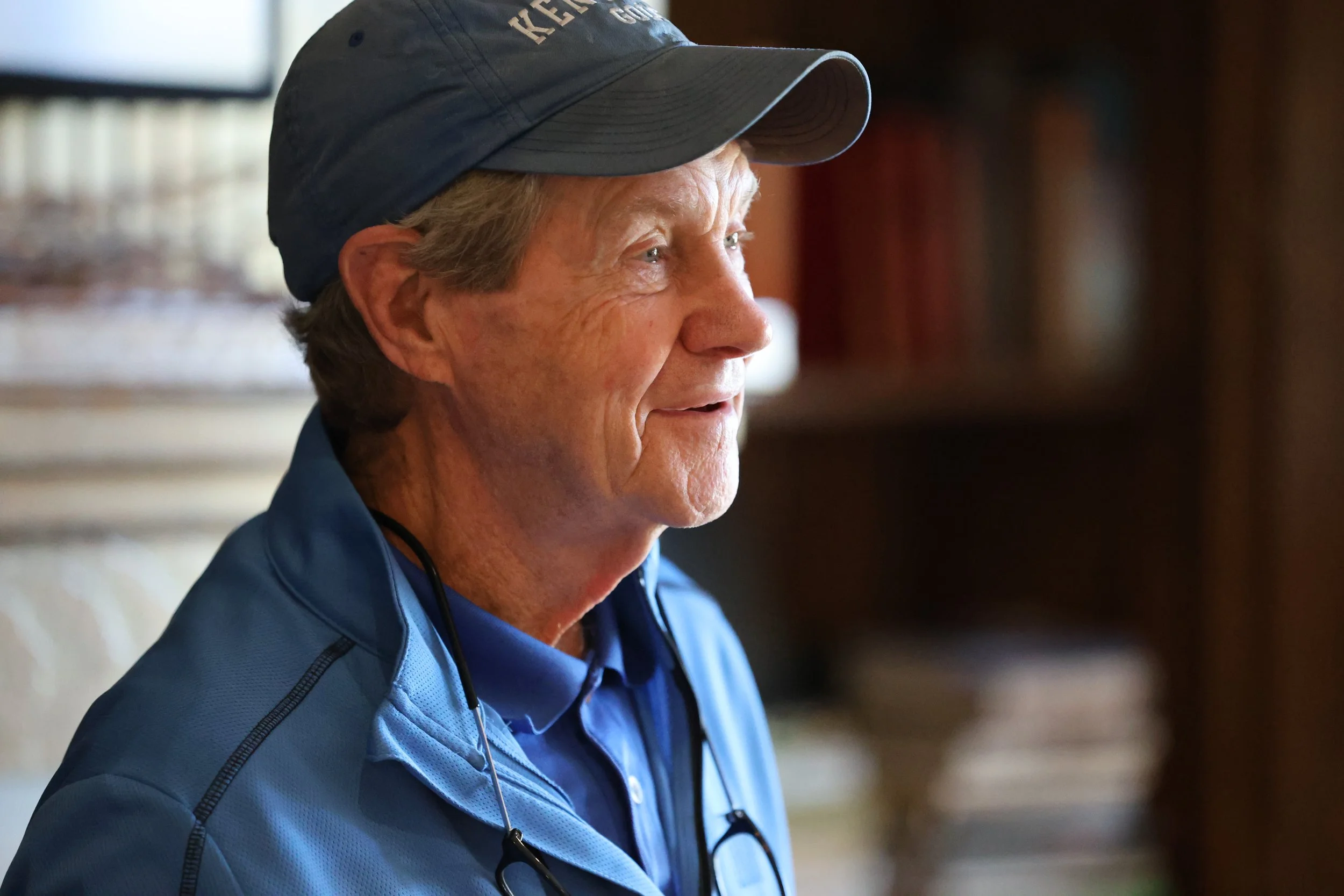 Profile of an elderly man wearing a blue jacket, blue shirt, and a cap, with glasses hanging around his neck, indoors.