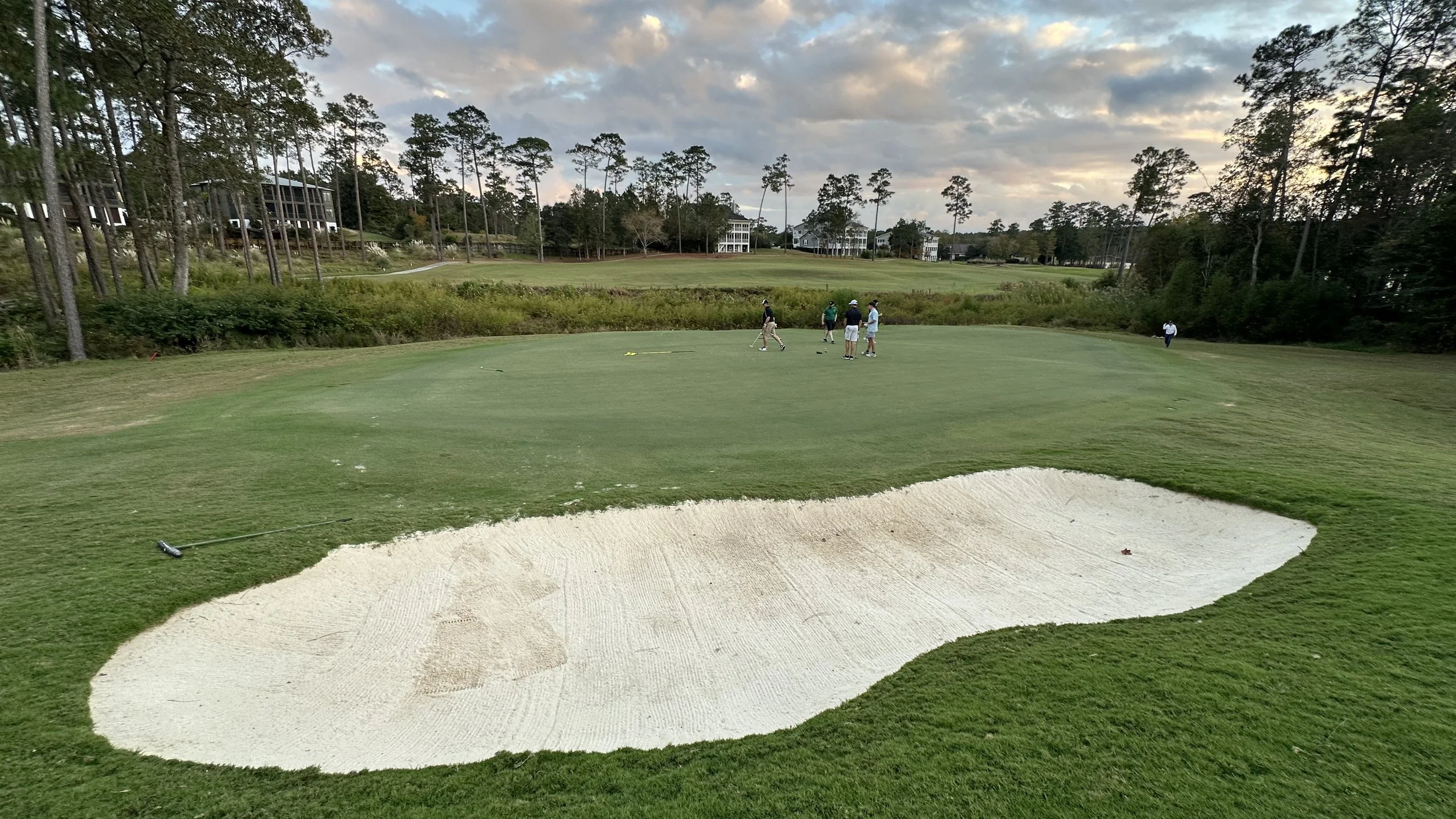 A golf course green with a sand bunker in the foreground and several people playing golf in the background, surrounded by trees and residential buildings.