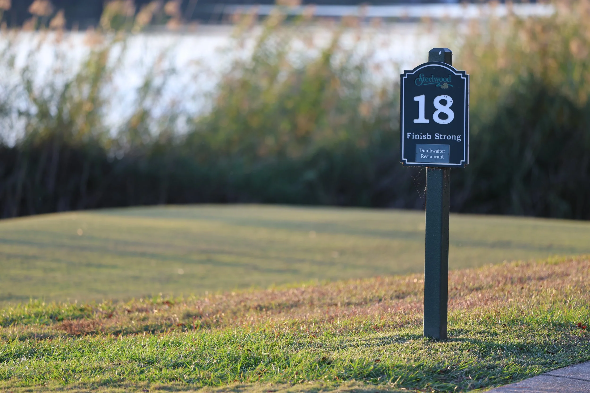 Golf course with a sign that reads 'Steelwood 18 Finish Strong' in the foreground, with trees and a water feature in the background.