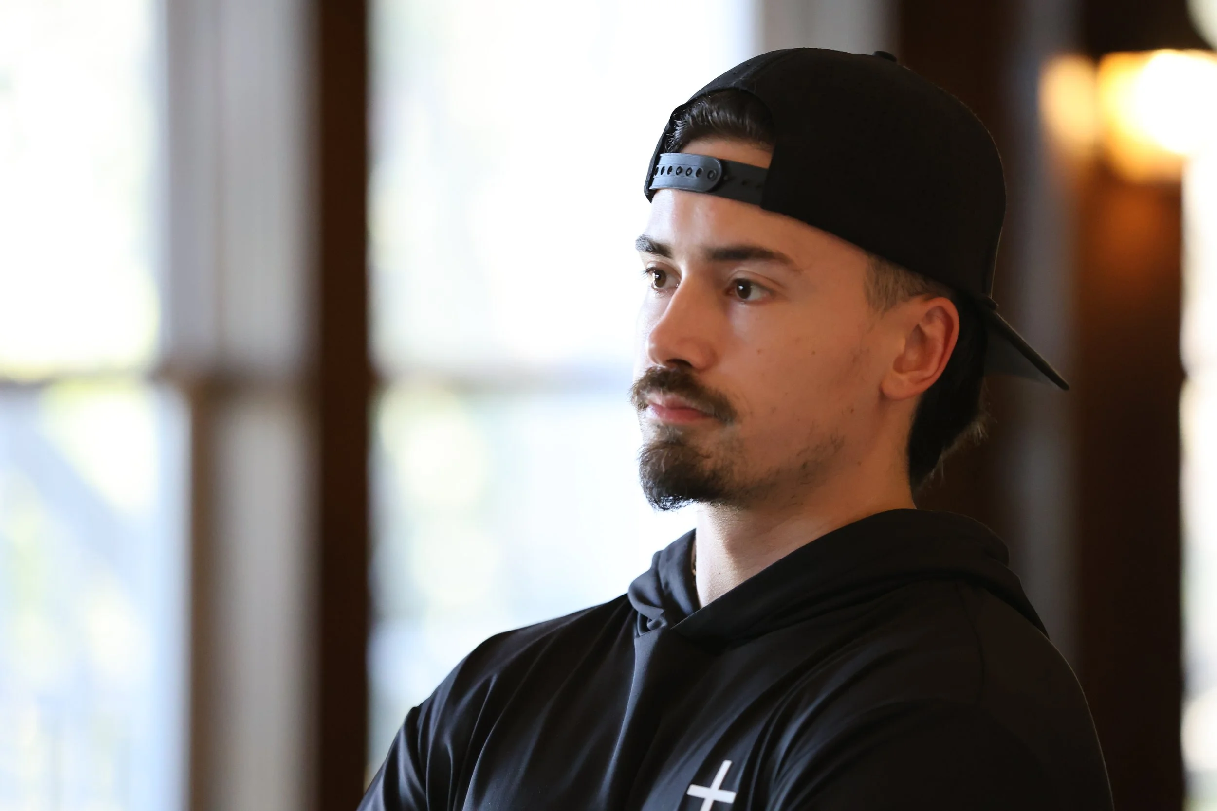 A young man with a mustache and goatee, wearing a black baseball cap backwards and a black hoodie, looking to the side indoors with blurred windows in the background.