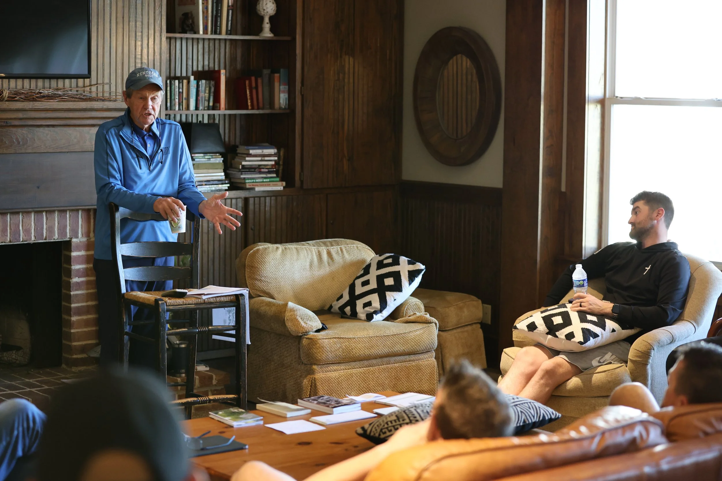An older man in a blue jacket and cap is speaking to a group of seated people in a cozy living room. One man is sitting on an armchair holding a water bottle, listening attentively, while others are relaxed on a couch. The room has wooden paneling, a