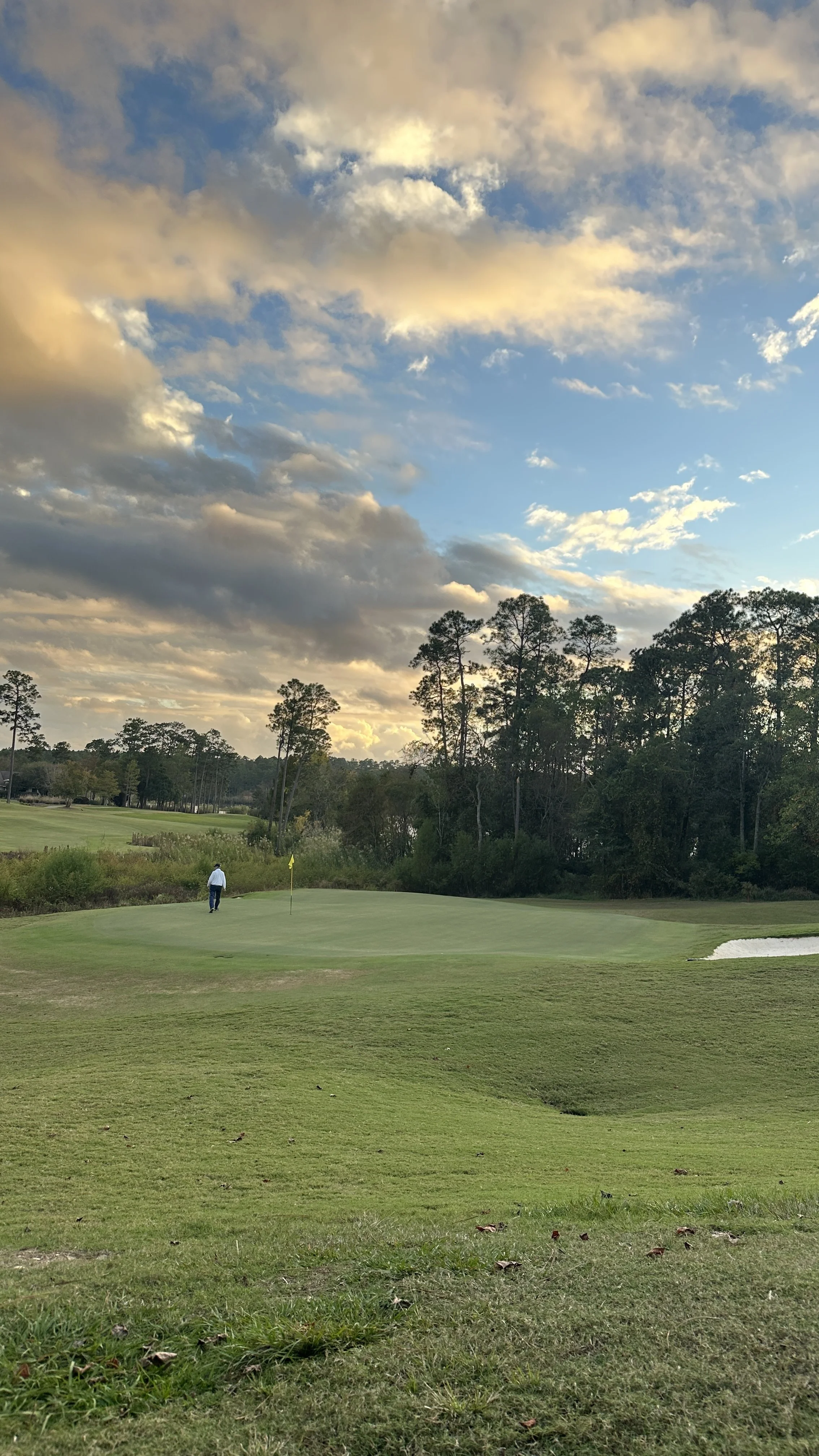 A person walking on a golf course near a green, with trees, a water hazard, and a partly cloudy sky during sunset.