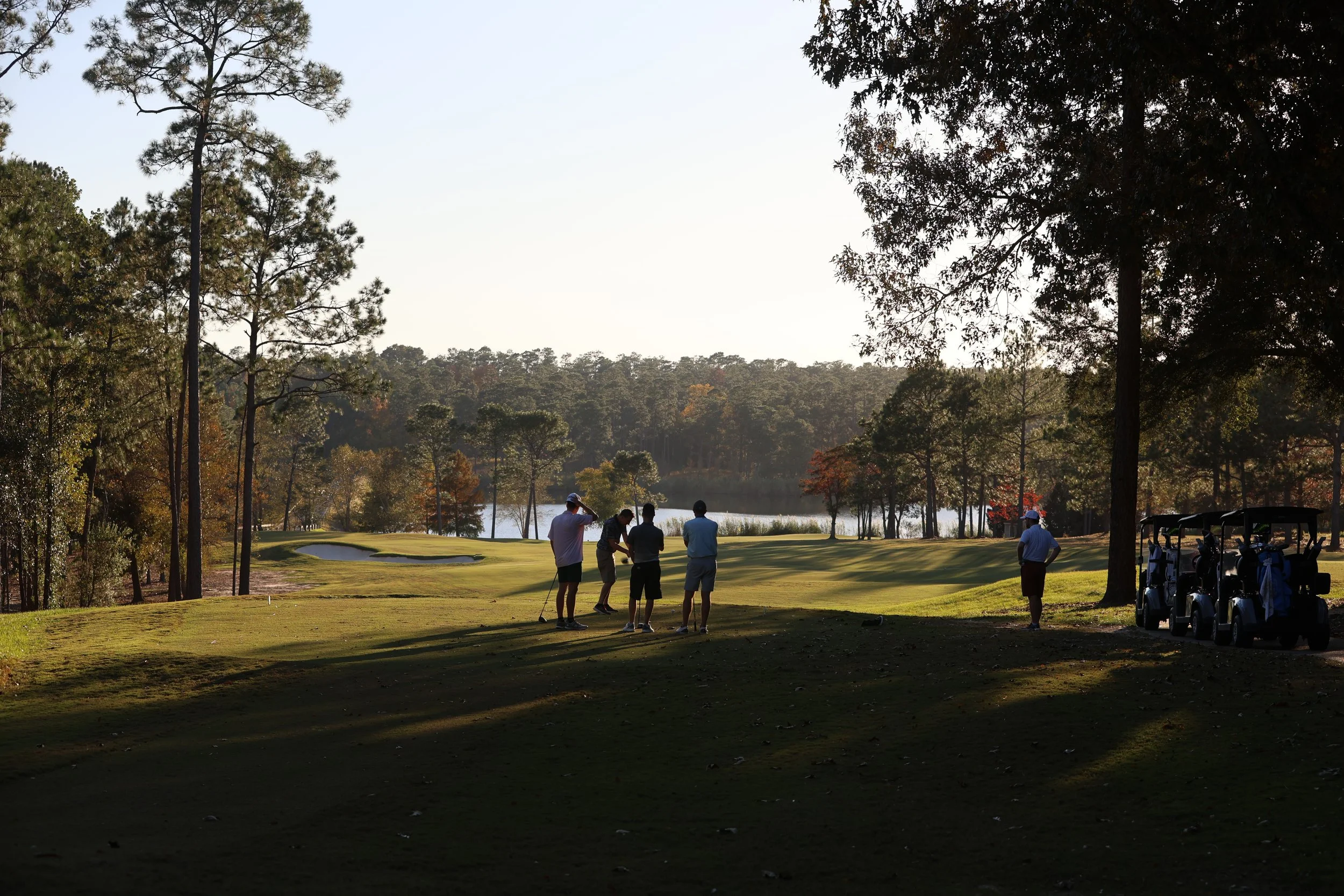 Golfers on a golf course playing near a pond during sunset, with trees and golf carts nearby.