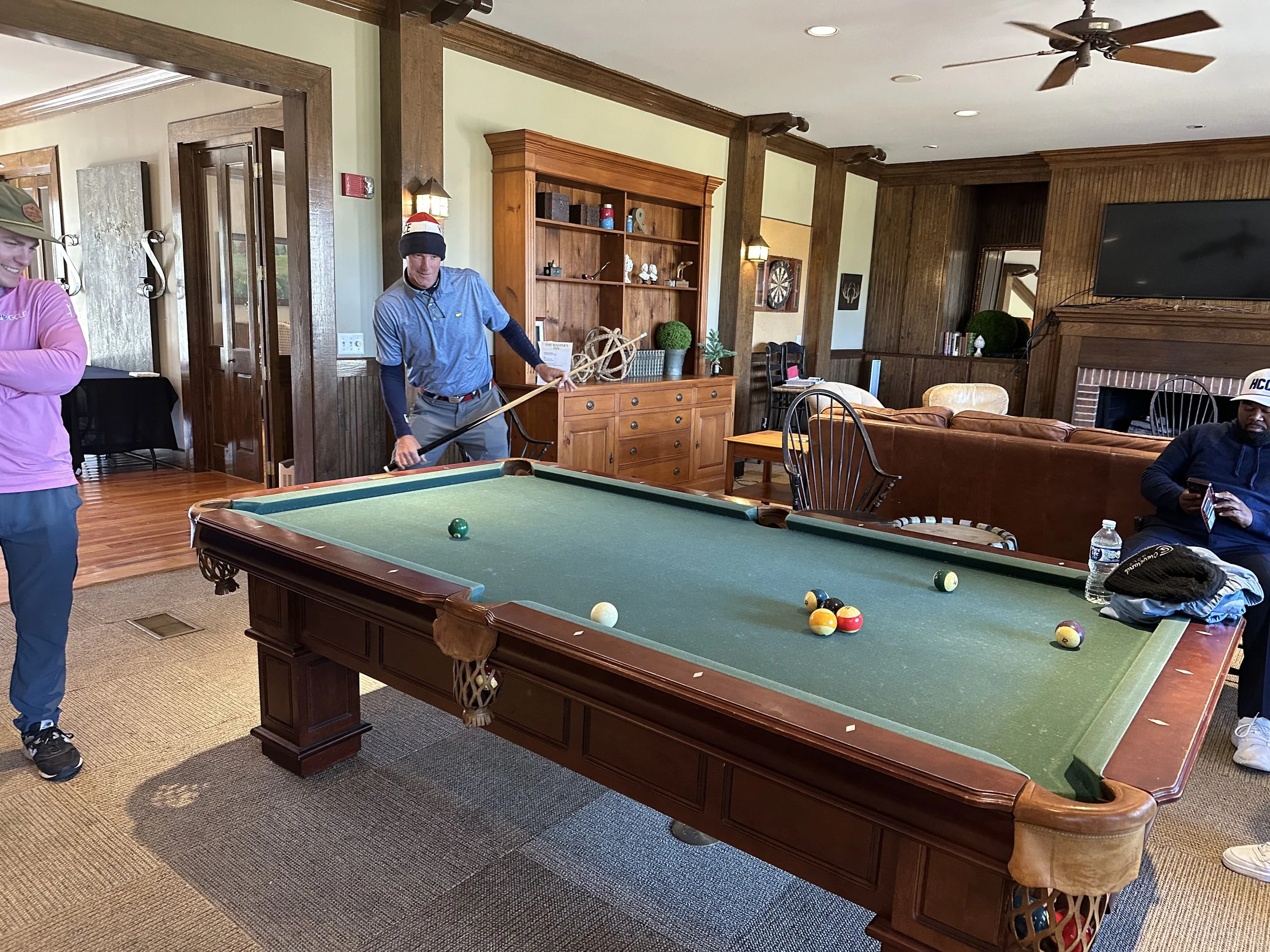People playing pool in a cozy living room with wood paneling and a fireplace.