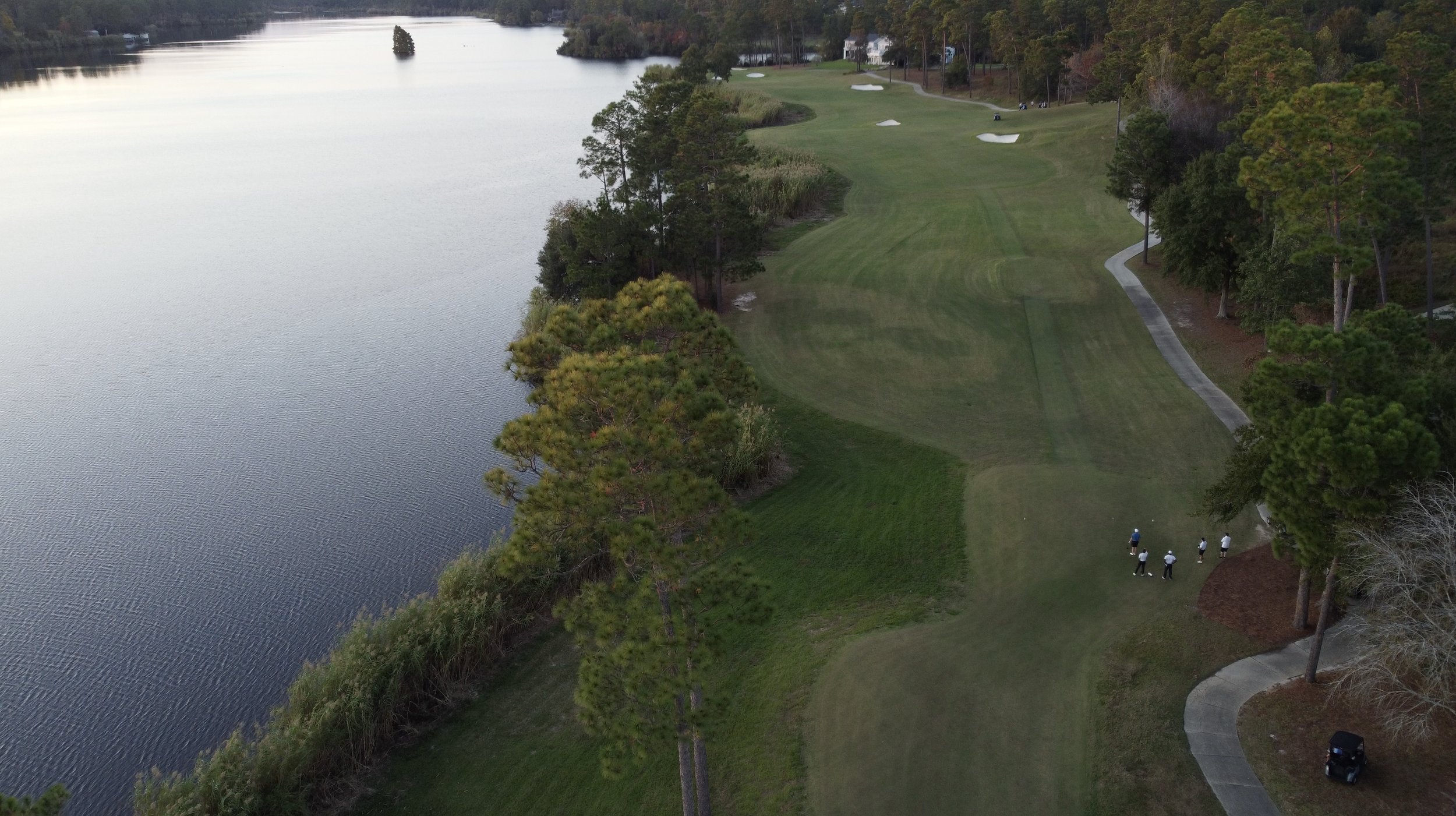 Aerial view of a golf course next to a large body of water, with four golfers walking on the green, surrounded by trees and a cart path.