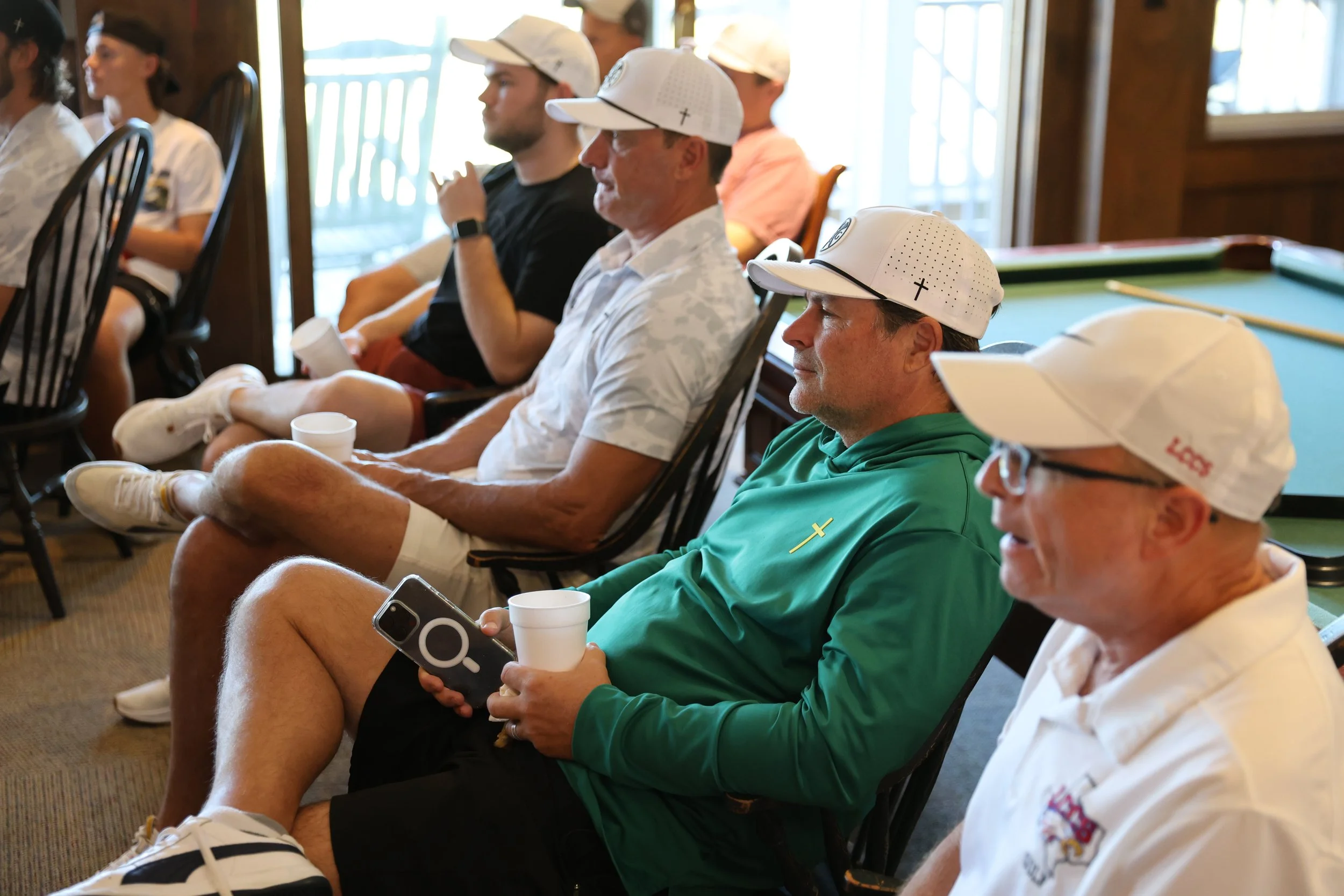 Group of people sitting indoors, wearing golf caps, some holding cups, with a pool table in the background.
