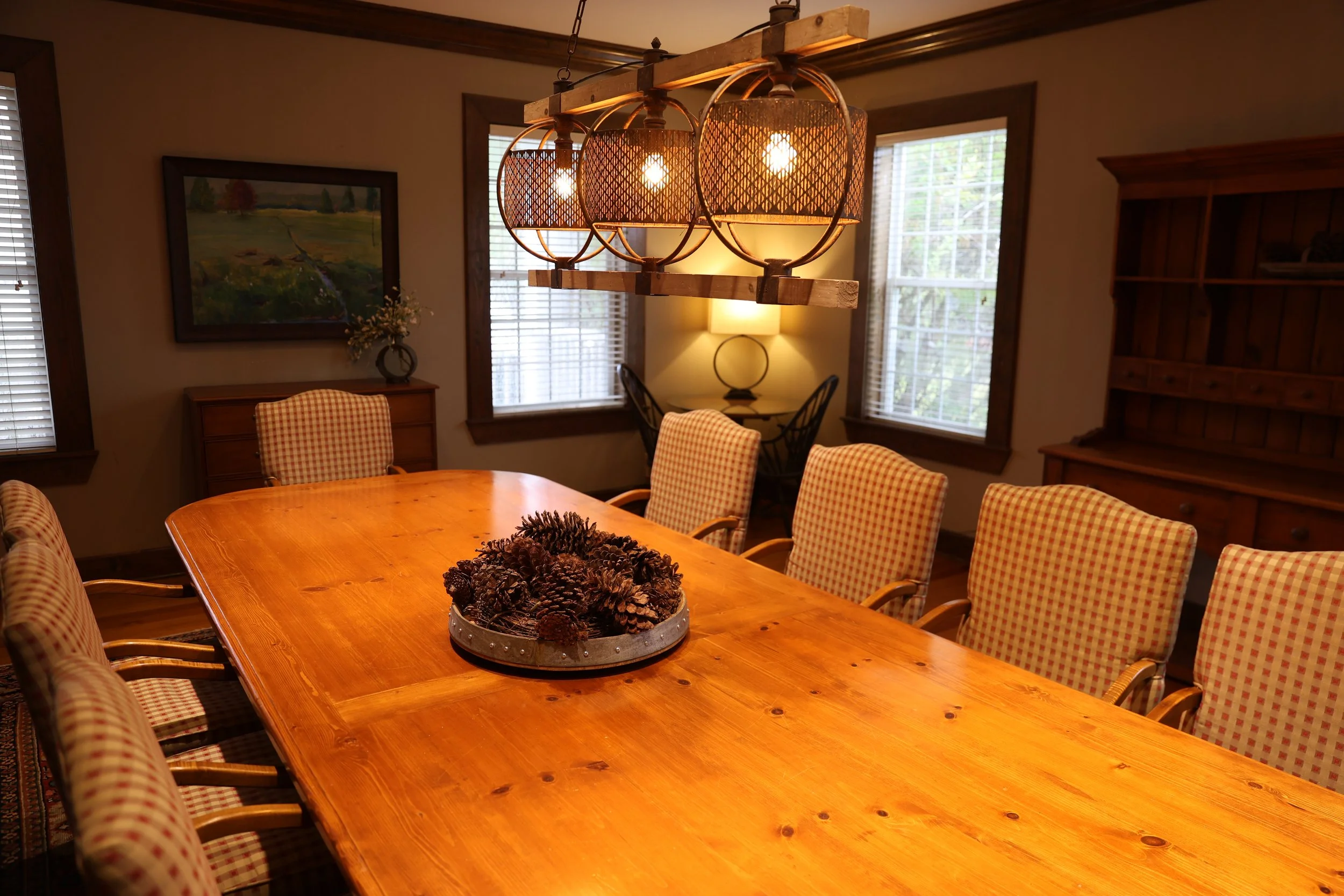 A  room with a large wooden table surrounded by upholstered chairs with checkered fabric. There is a decorative centerpiece with pinecones on the table. A modern hanging light fixture with multiple bulbs and woven shades hangs above the table. The ro