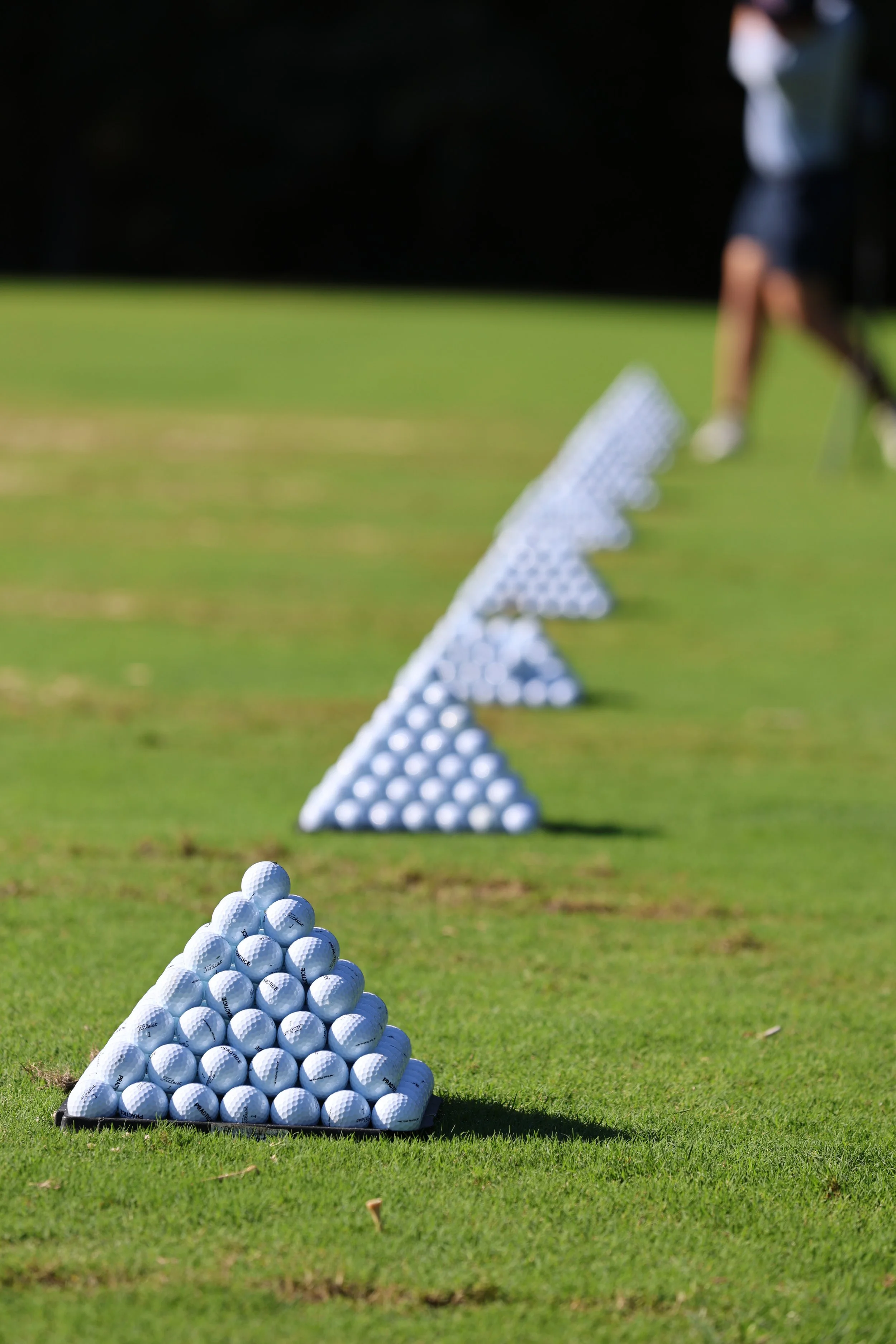 Golf practice area with golf balls arranged in a pyramid shape on the ground, and more pyramids in the background leading towards a person in golf attire preparing to hit a ball.