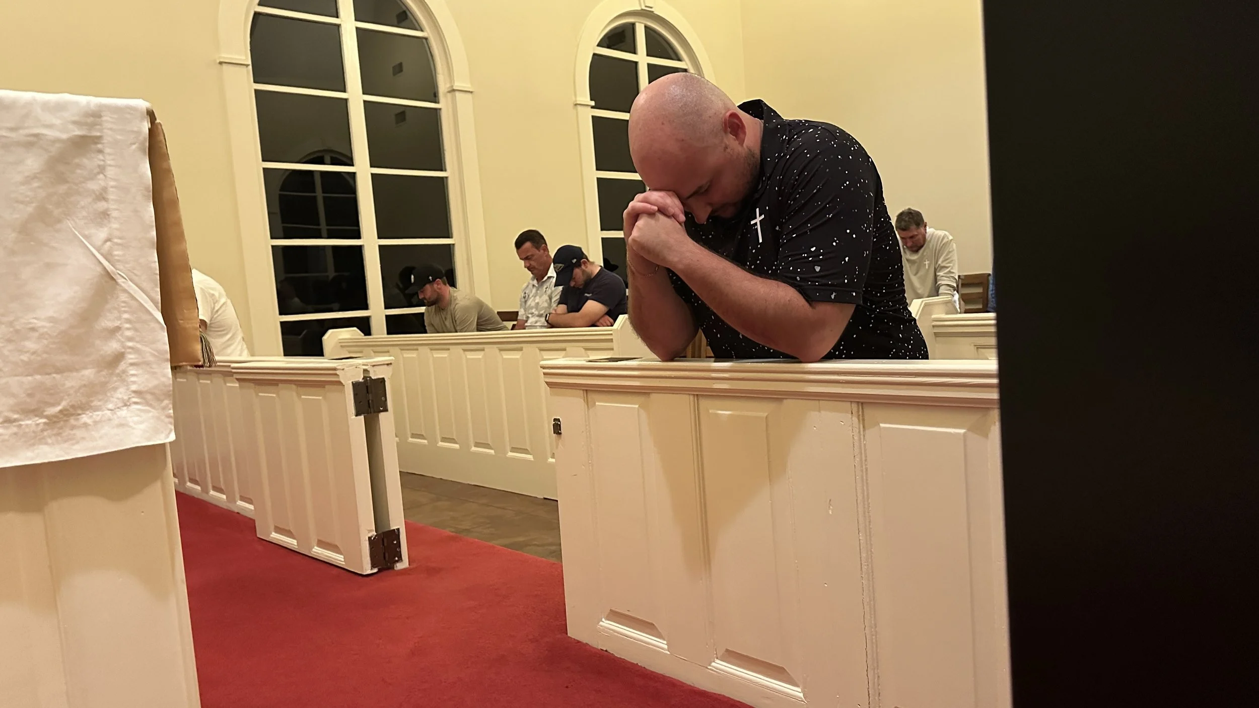 A man kneeling and praying in a church during a prayer service, with other people sitting in the pews behind him, some with heads bowed and eyes closed.