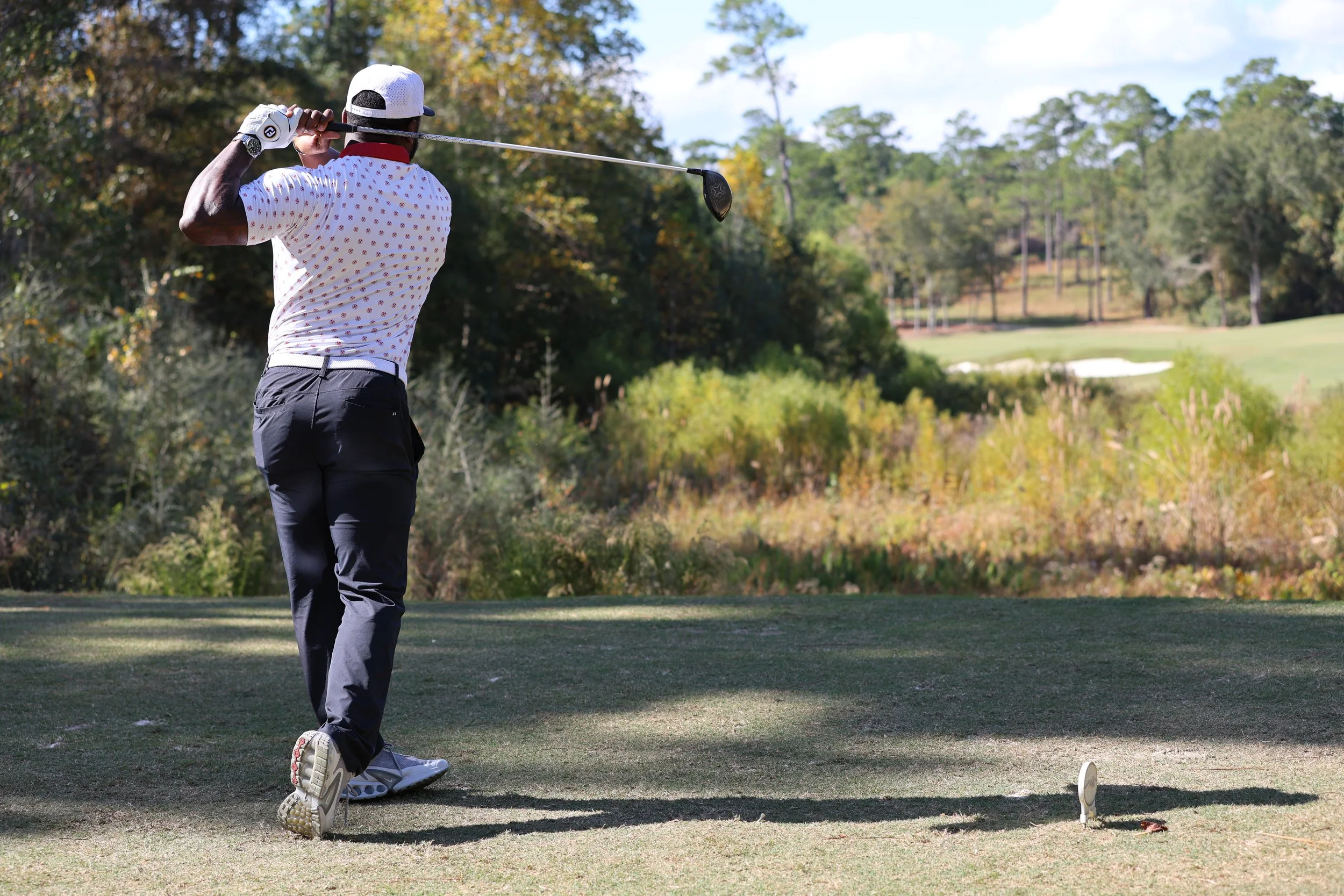 Man playing golf, taking a shot on a golf course with trees and a bunker in the background.