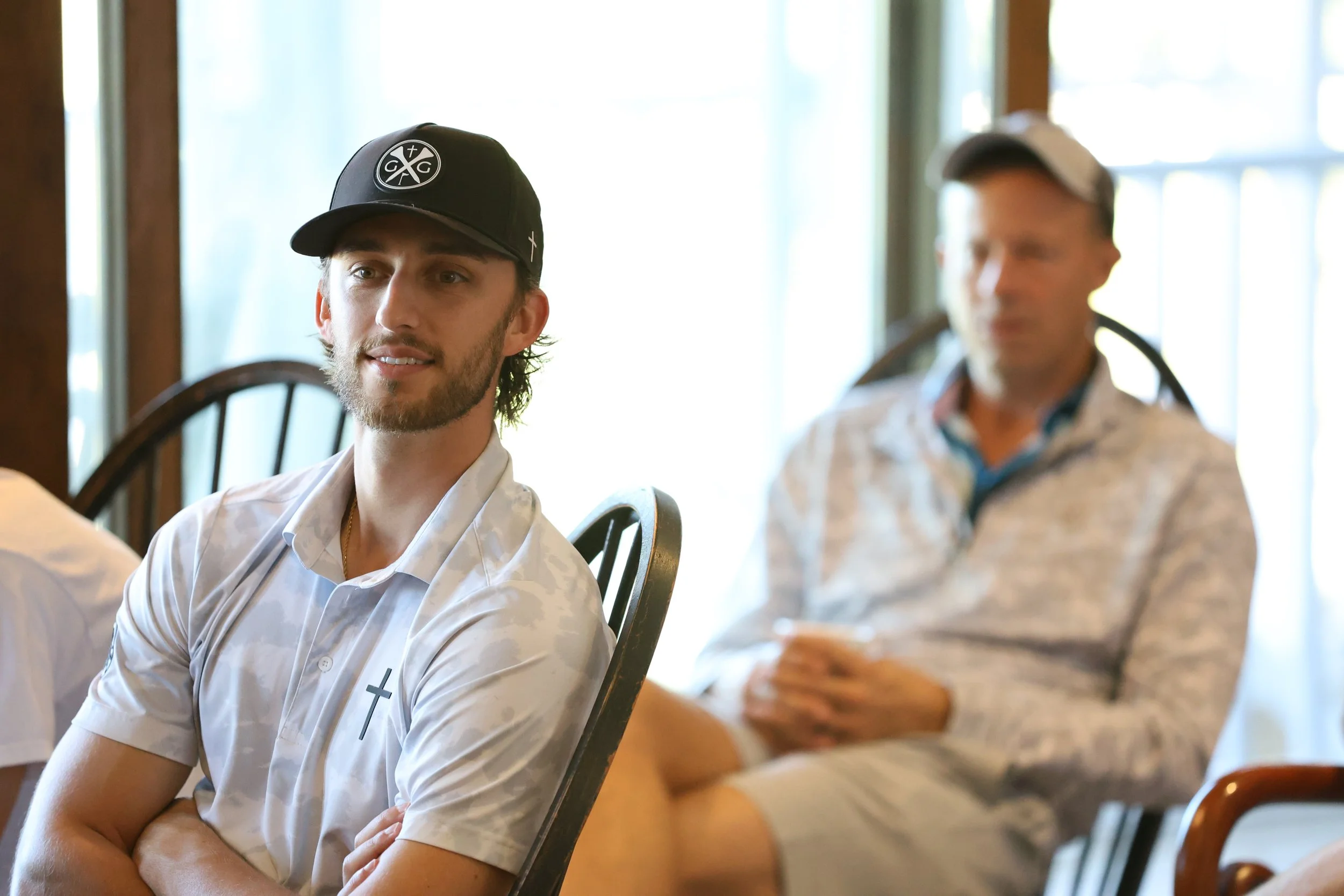 A young man with a beard and long hair, wearing a black baseball cap, white shirt with a cross on it, sitting on a chair indoors near a window, smiling slightly, with another man blurred in the background.