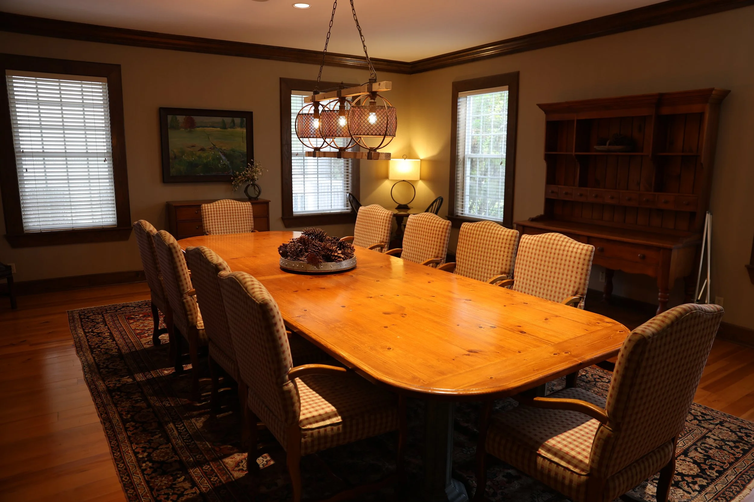 A dining room with a large wooden table surrounded by ten plaid upholstered chairs, a chandelier hanging above, windows with blinds, a rug under the table, and a wooden hutch against the wall.
