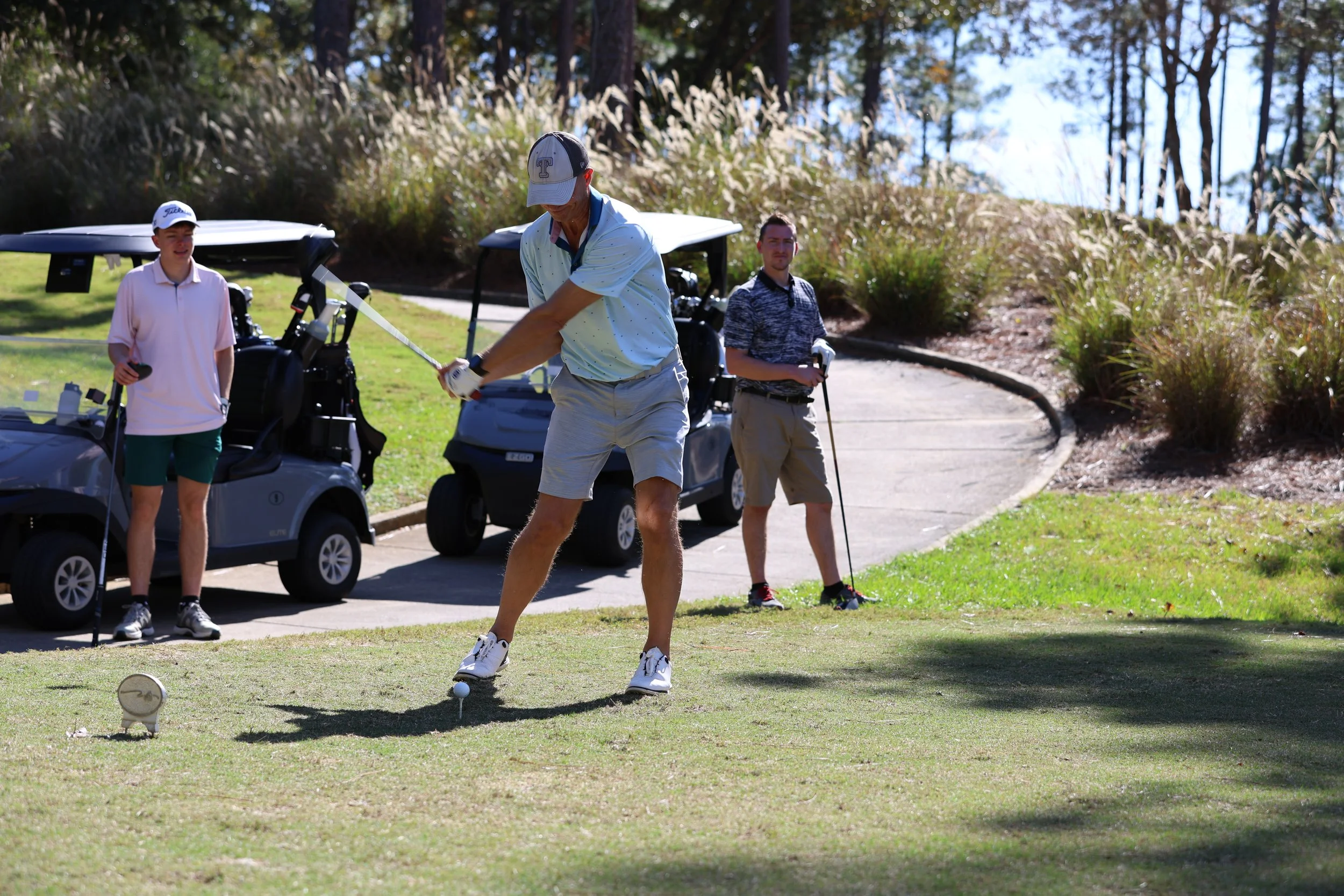 Three men playing golf at a golf course, with two parked golf carts behind them. One man is taking a swing while the others watch, surrounded by trees and sunny weather.