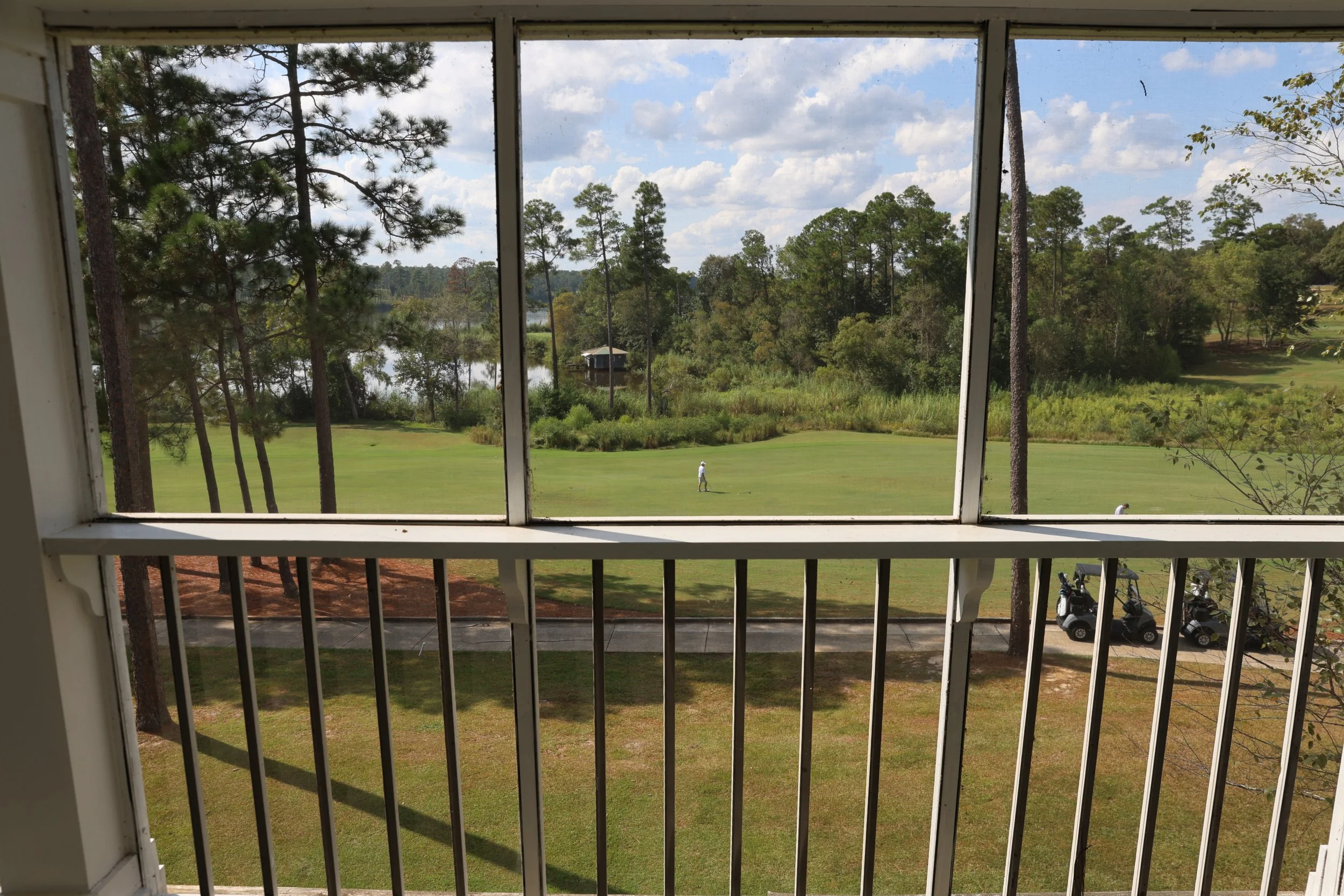 View of a golf course seen through a screened balcony window, with a golfer near the pond, surrounded by trees and golf carts parked nearby.