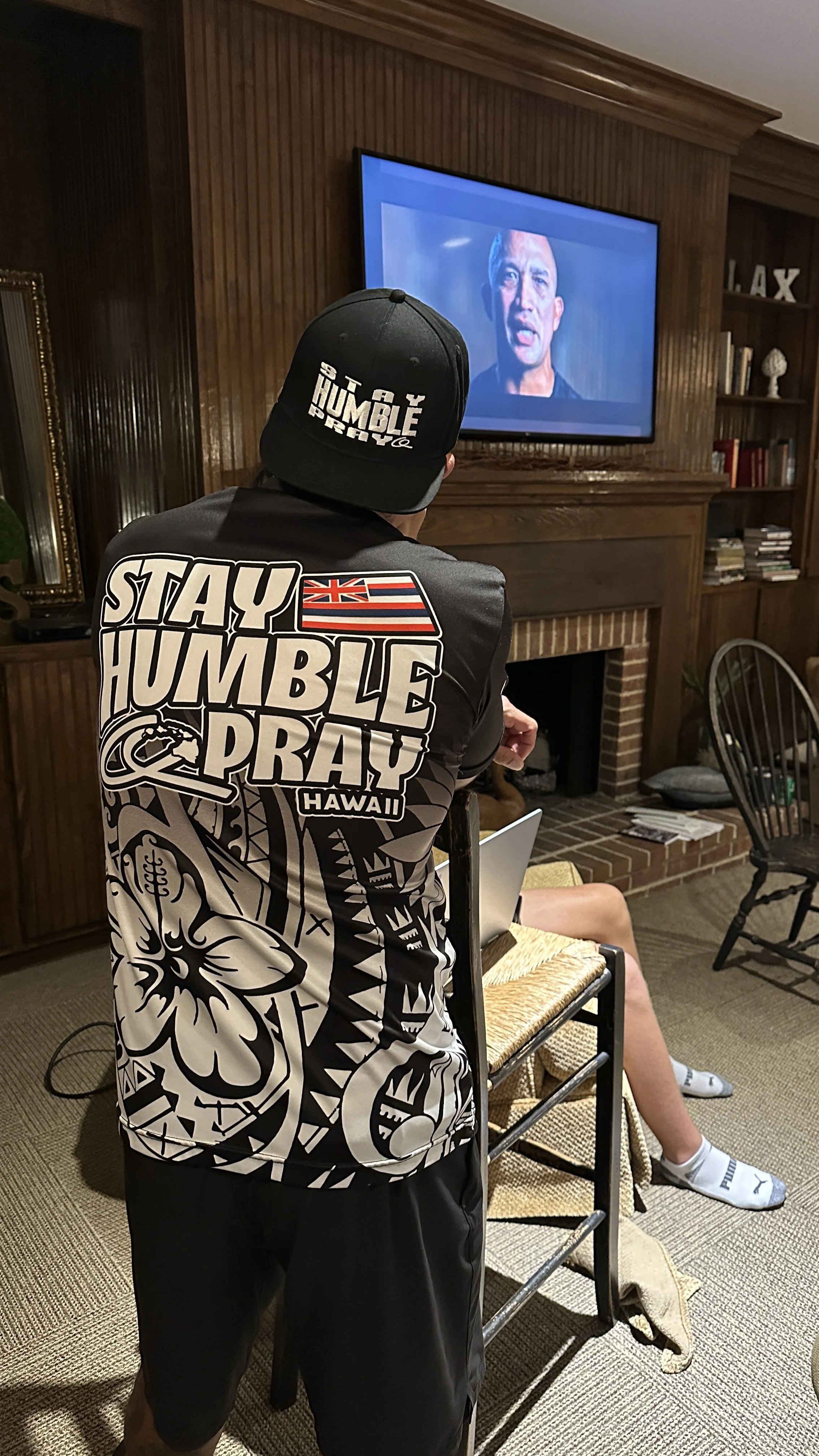 A man sits on a chair watching a television in a living room, wearing a black and white shirt with the words 'STAY HUMBLE PRAY' and a Hawaiian flag patch, along with a black baseball cap that says 'STAY HUMBLE'.