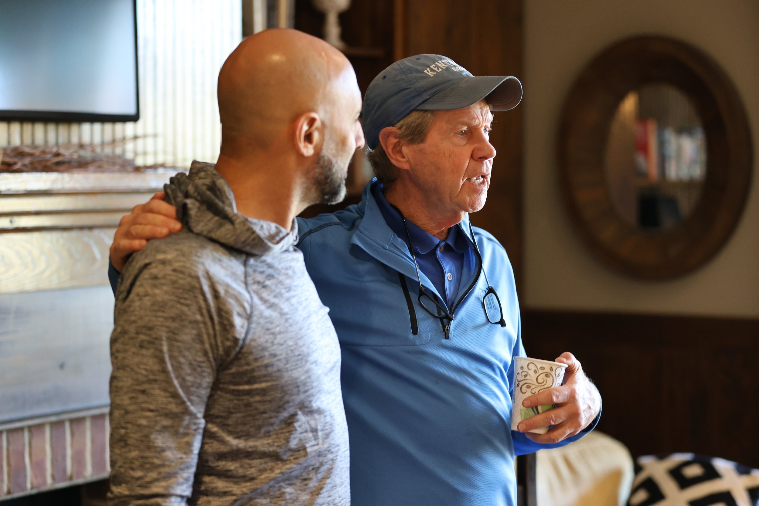 Two men stand close together indoors, one with his arm around the other's shoulder. The man on the right wears a blue jacket and cap, holding a mug, and appears to be speaking. The man on the left has a shaved head and beard, wearing a gray hoodie, a