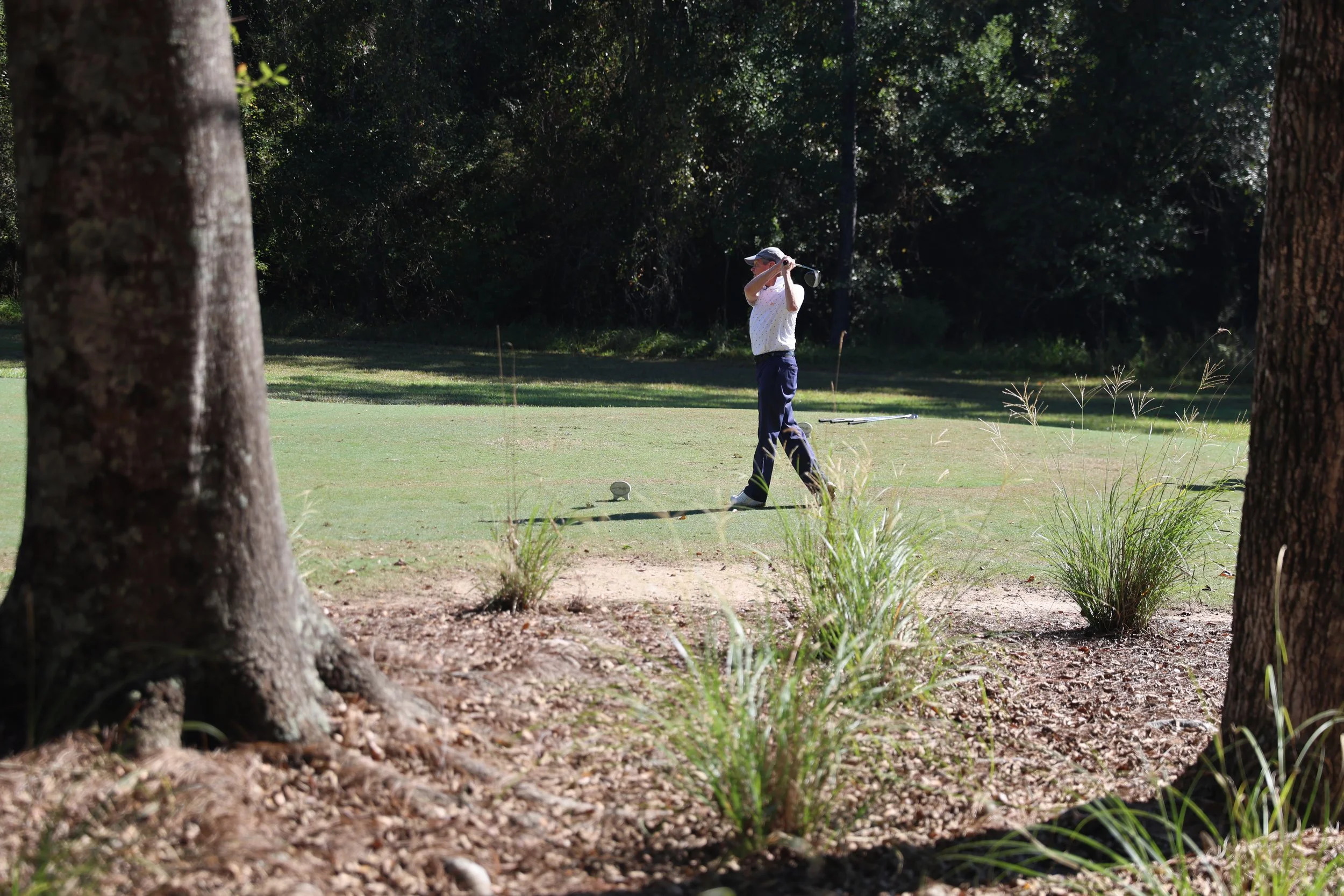 A person playing golf on a course, seen from behind through trees, preparing to hit a golf ball.