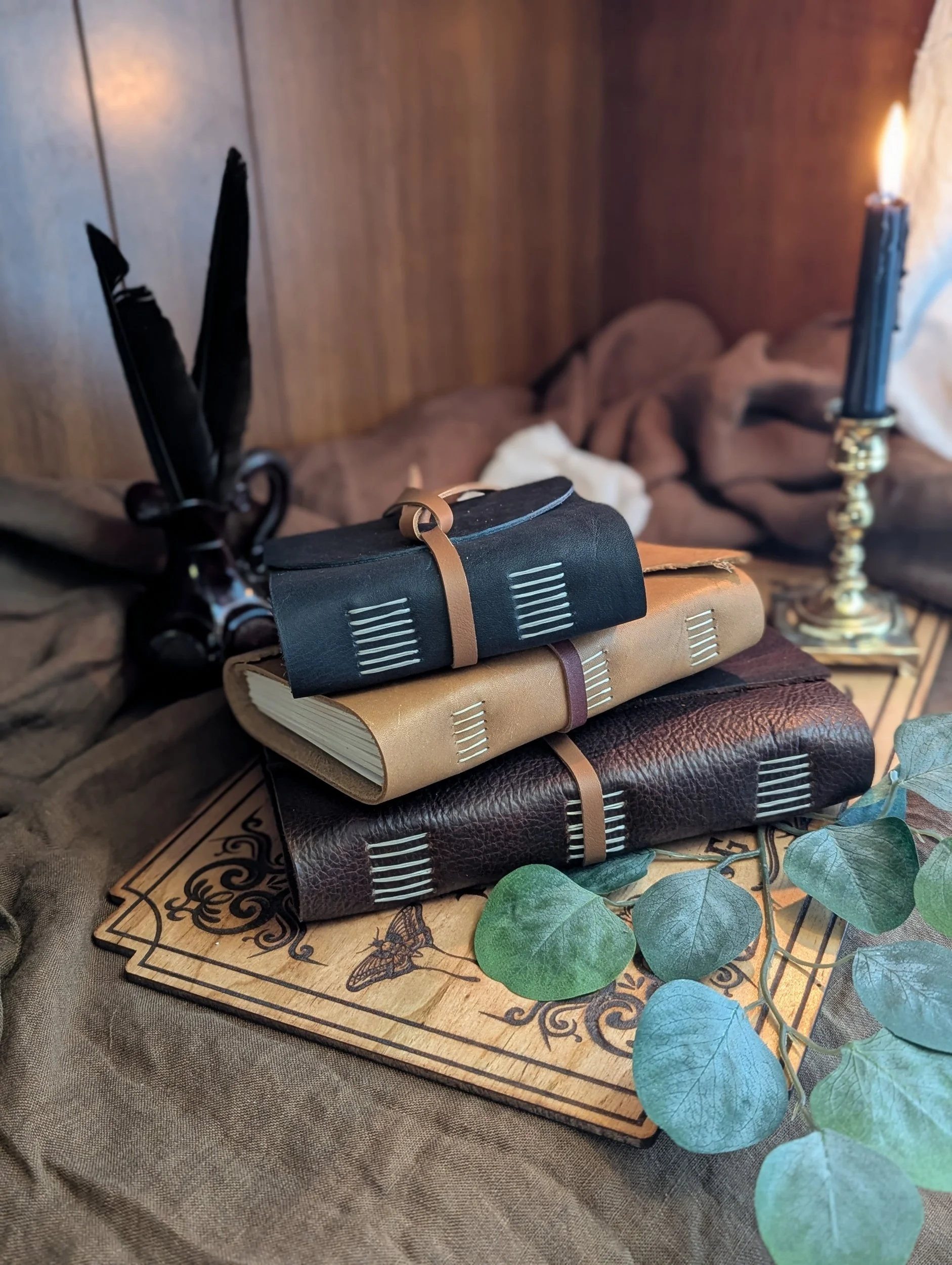 Stacked journals or notebooks with black, beige, and dark brown covers on a decorative wooden tray, surrounded by green eucalyptus leaves, on a textured fabric surface.