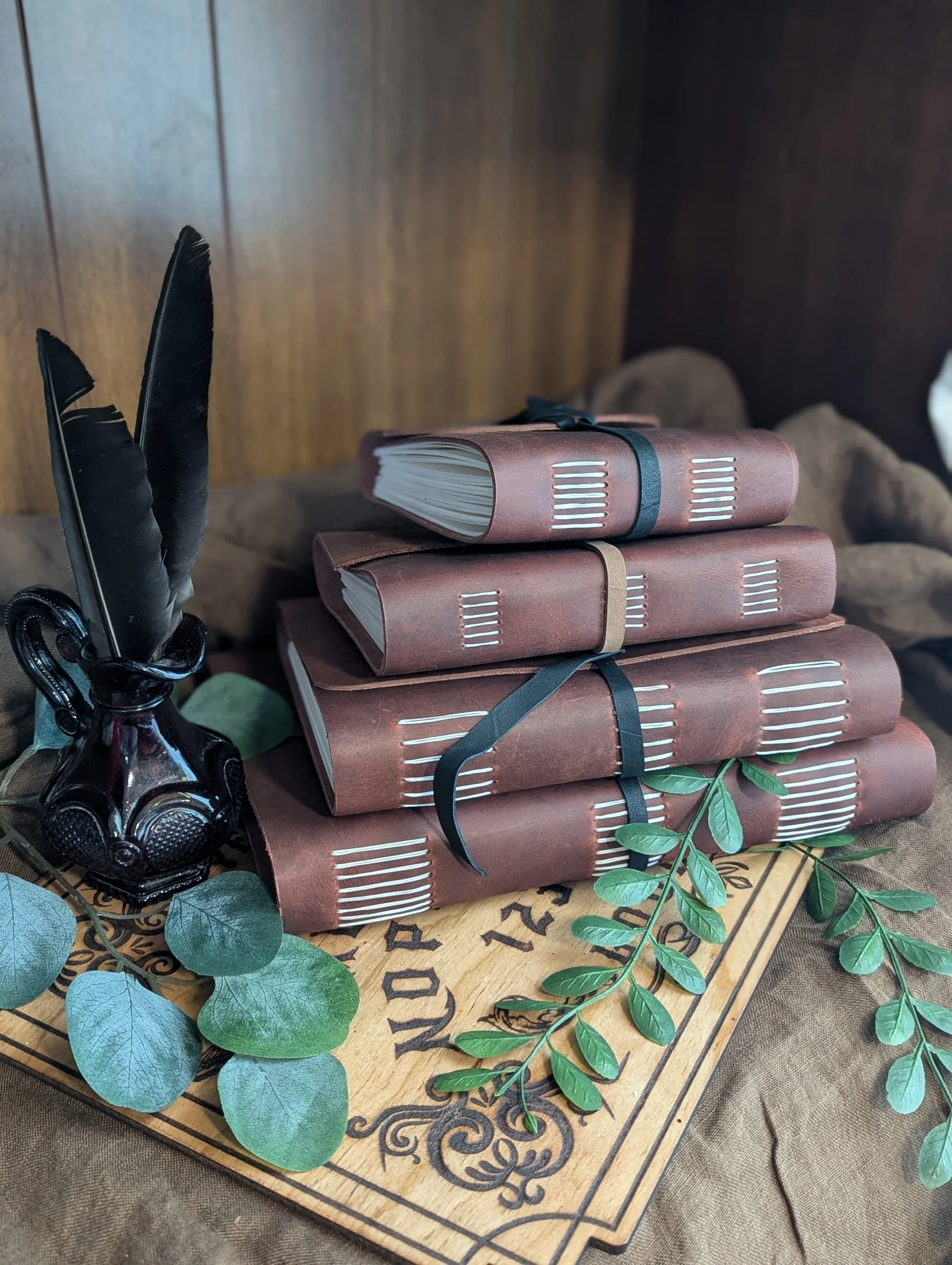 Stacked dark brown journals with black elastic bands, green leaves, and black feathers in a glass container on a decorative wooden board.