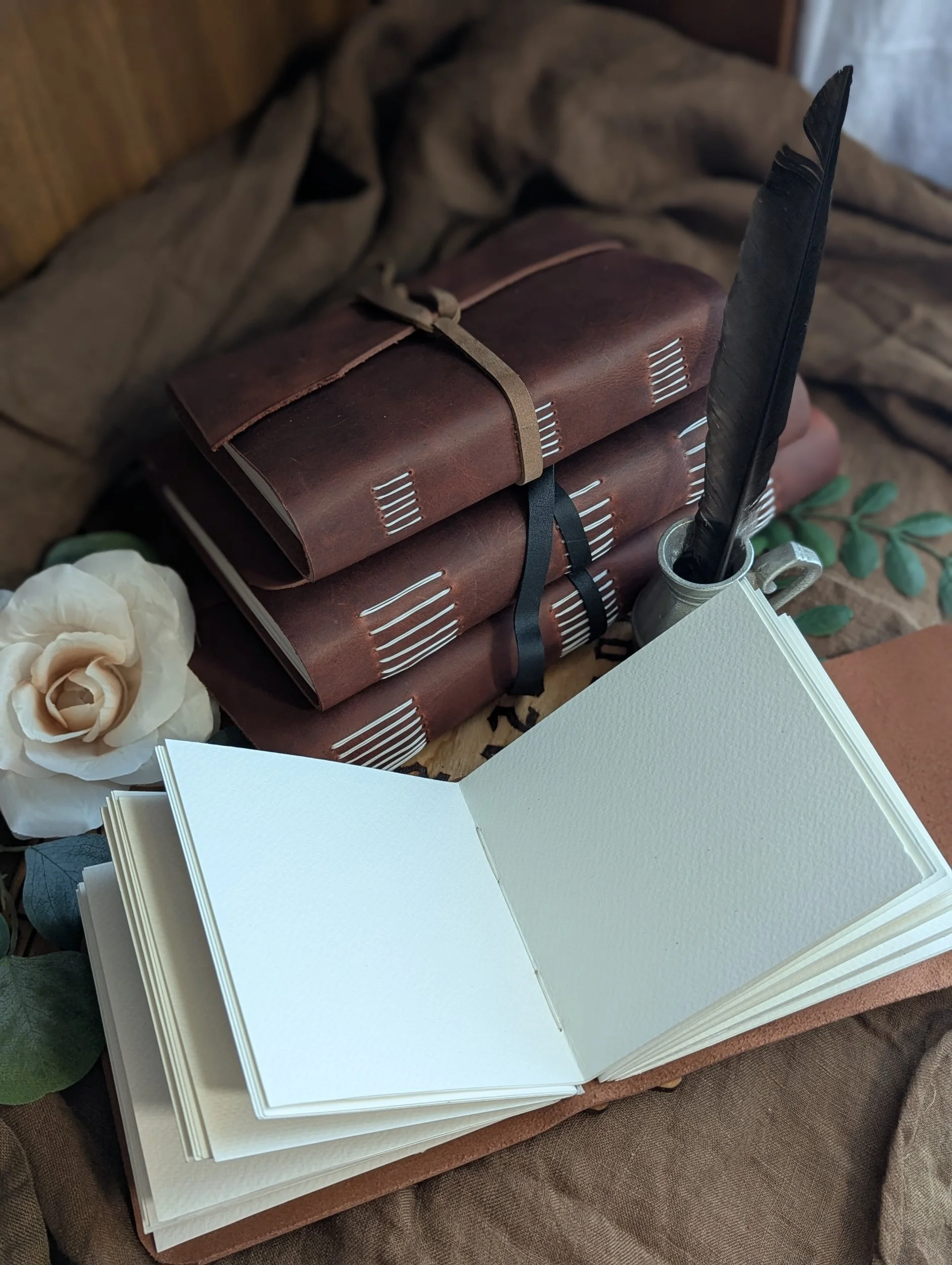 Open blank sketchbooks or watercolor pads stacked on a table, with a quill pen in an inkwell, surrounded by decorative leaves and a cream-colored flower.