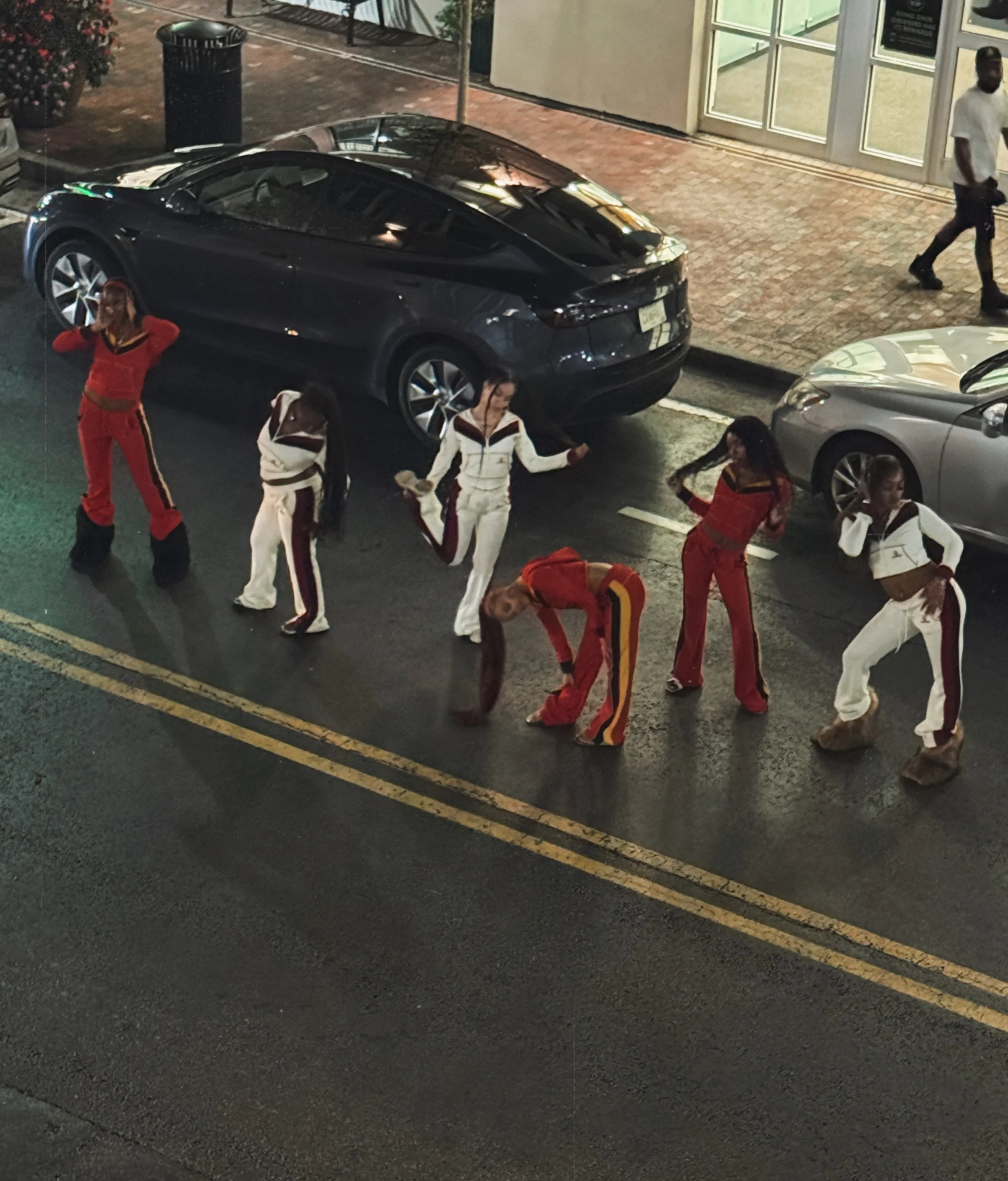 Six people in colorful outfits dancing on a city street at night, with parked cars and a building with glass windows in the background.