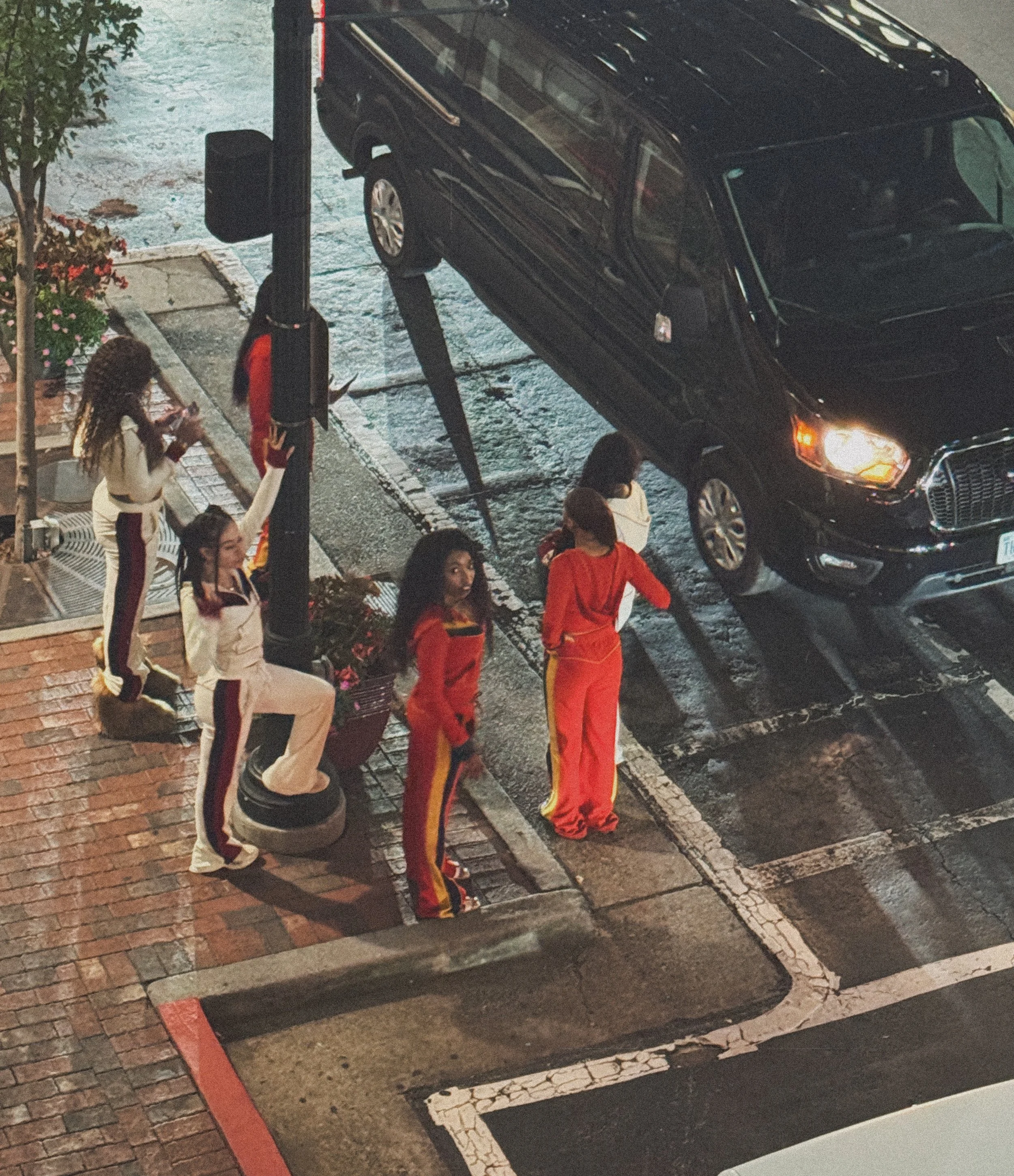 Five women in athletic wear, some standing and some sitting on tires, next to a parked black SUV on a city street at night, with some women engaging with their phones.