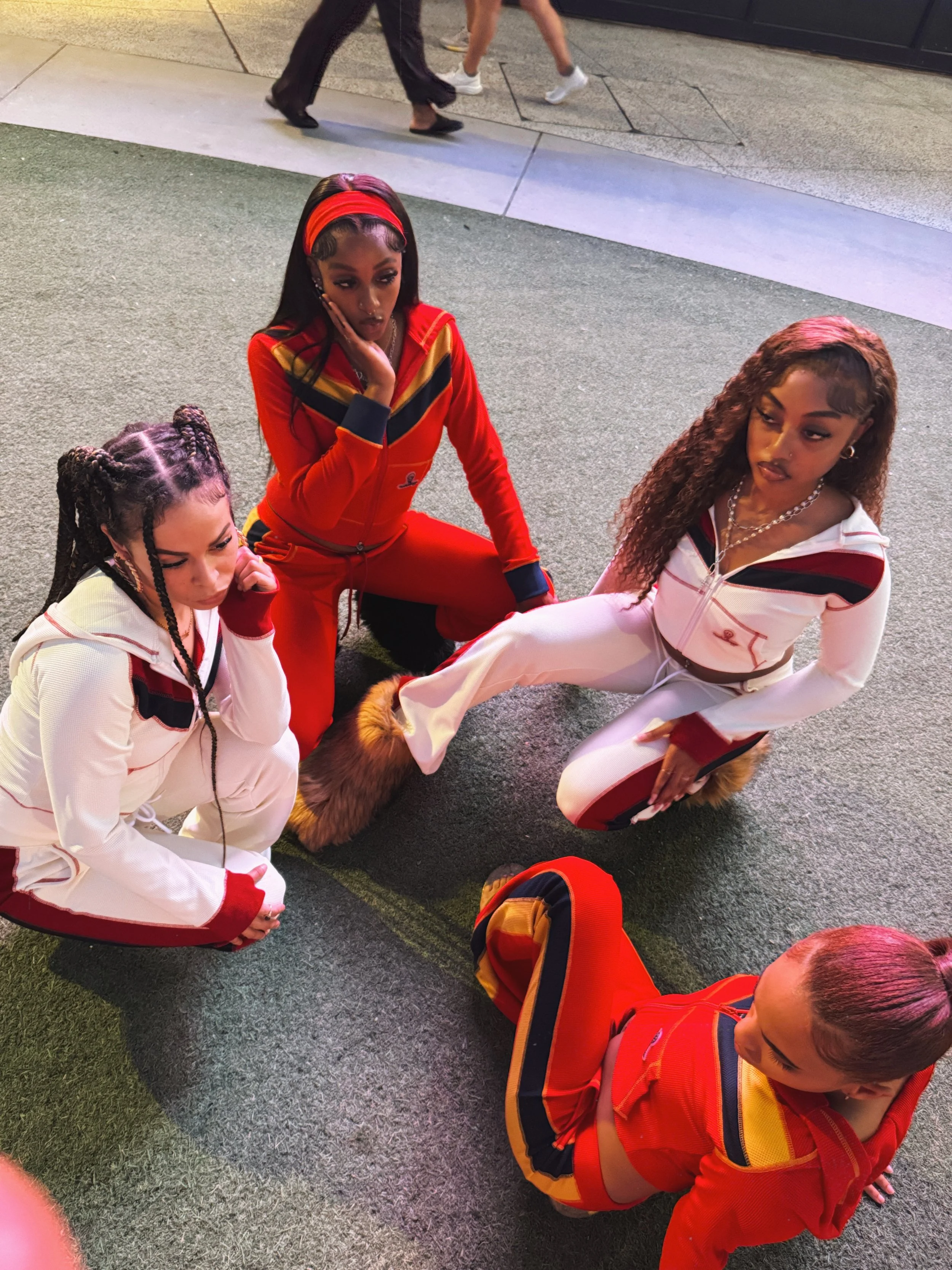 Four women dressed in sporty outfits sitting and kneeling on a textured gray surface outside, with two people walking in the background.