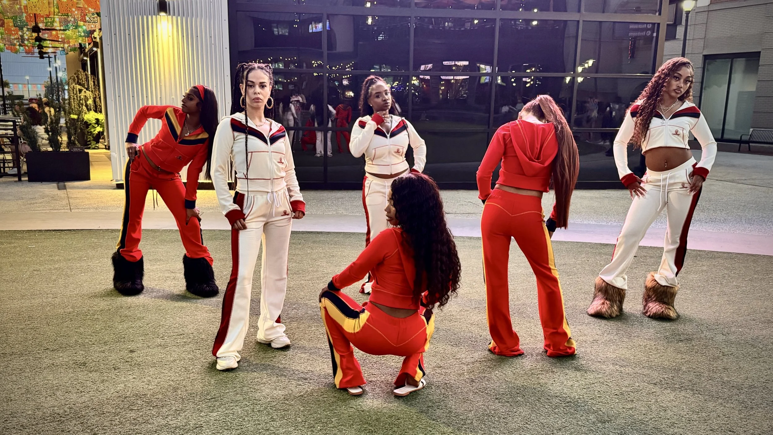 Six women dressed in colorful sporty outfits, with some wearing furry boots, standing on a grassy area in front of a glass building at night.