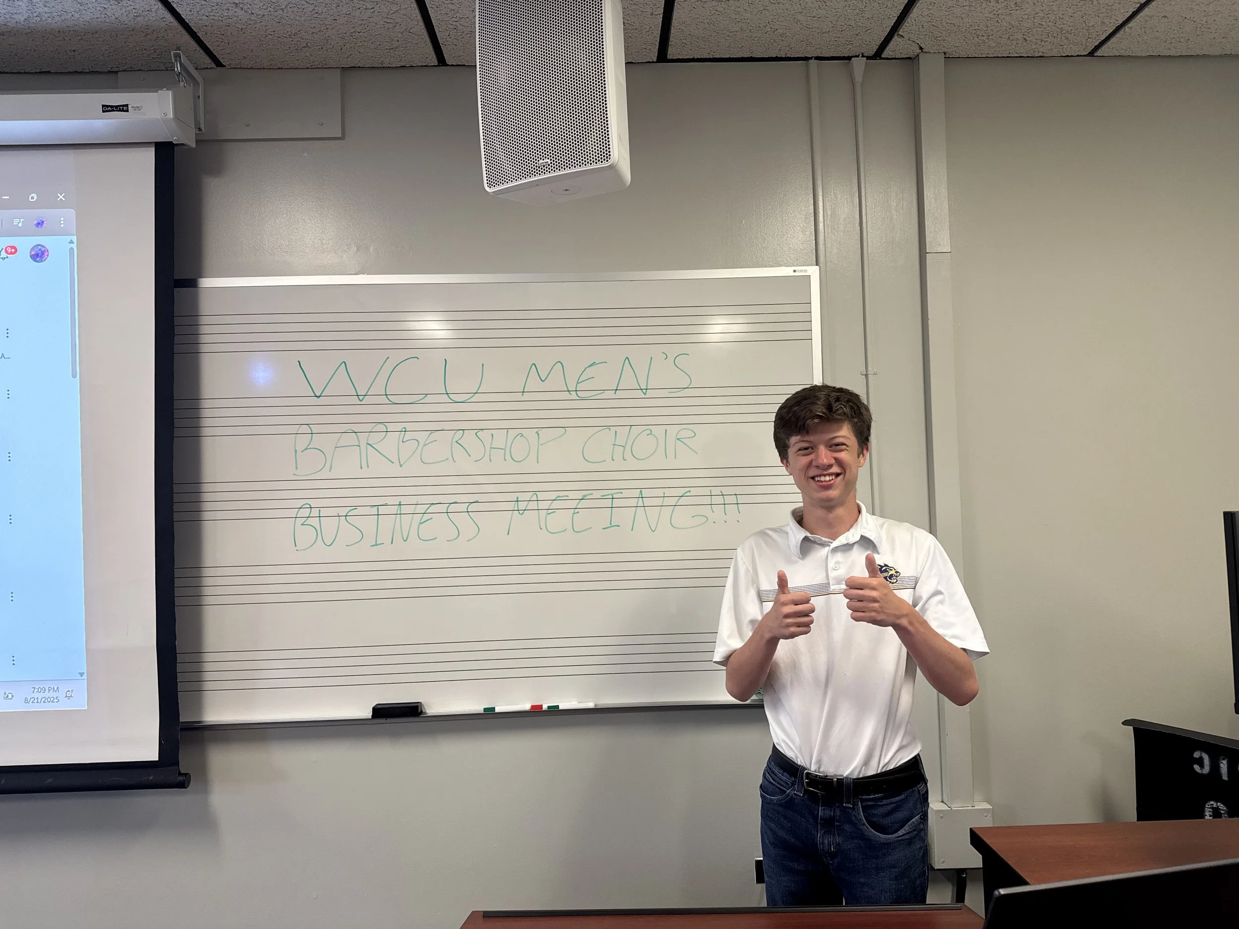 A young man standing in front of a whiteboard with green writing that says 'WCU MEN'S BARBERSHOP CHOIR BUSINESS MEETING!!!', smiling and giving two thumbs up, in a classroom or lecture room setting.