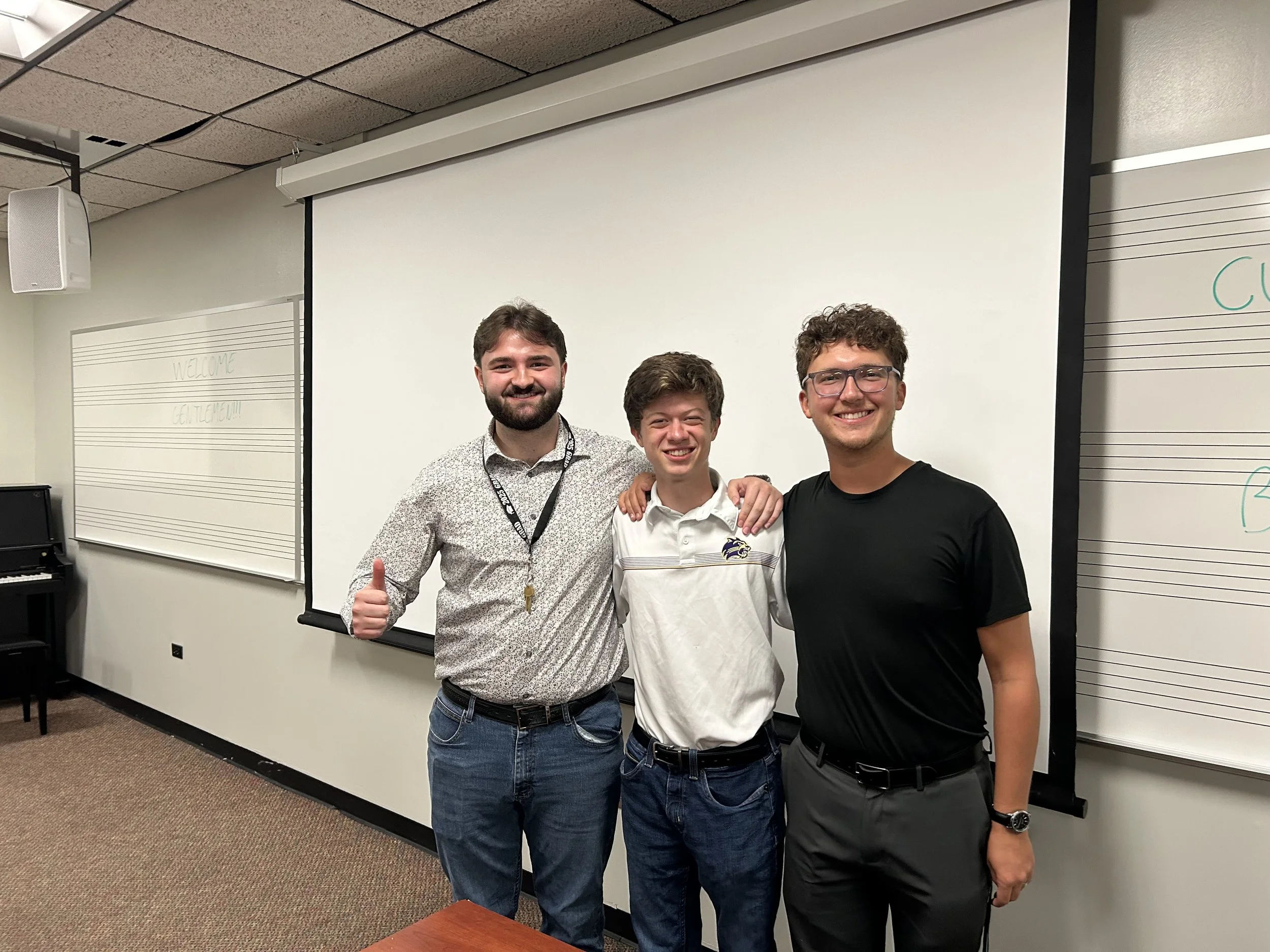 Three young men smiling and posing for a photo in a classroom with music staff lines on whiteboards behind them.