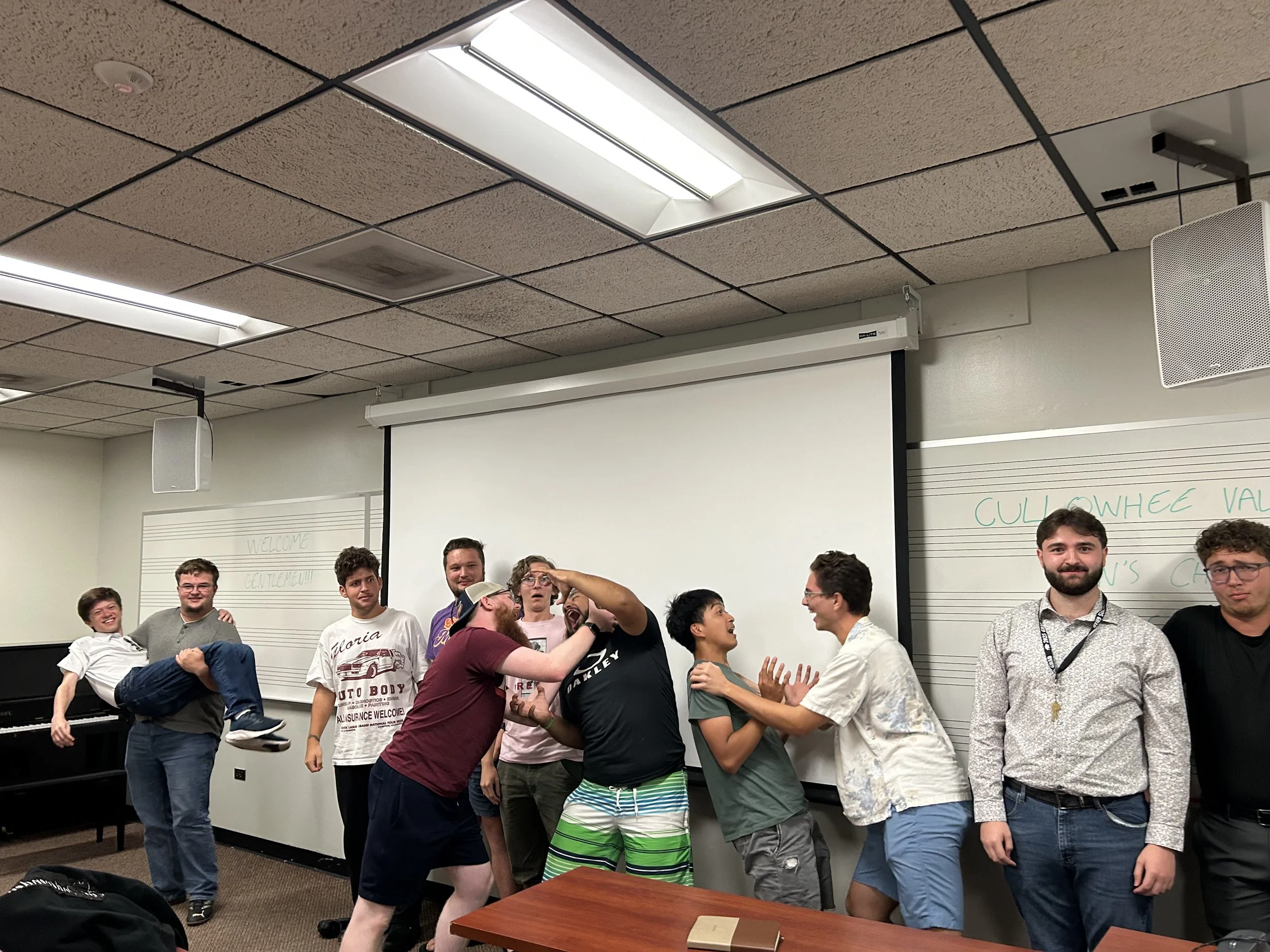 A group of students in a classroom engaging in playful and humorous interactions, with some pretending to physically fight and others smiling or laughing, standing in front of a whiteboard and a projector screen.
