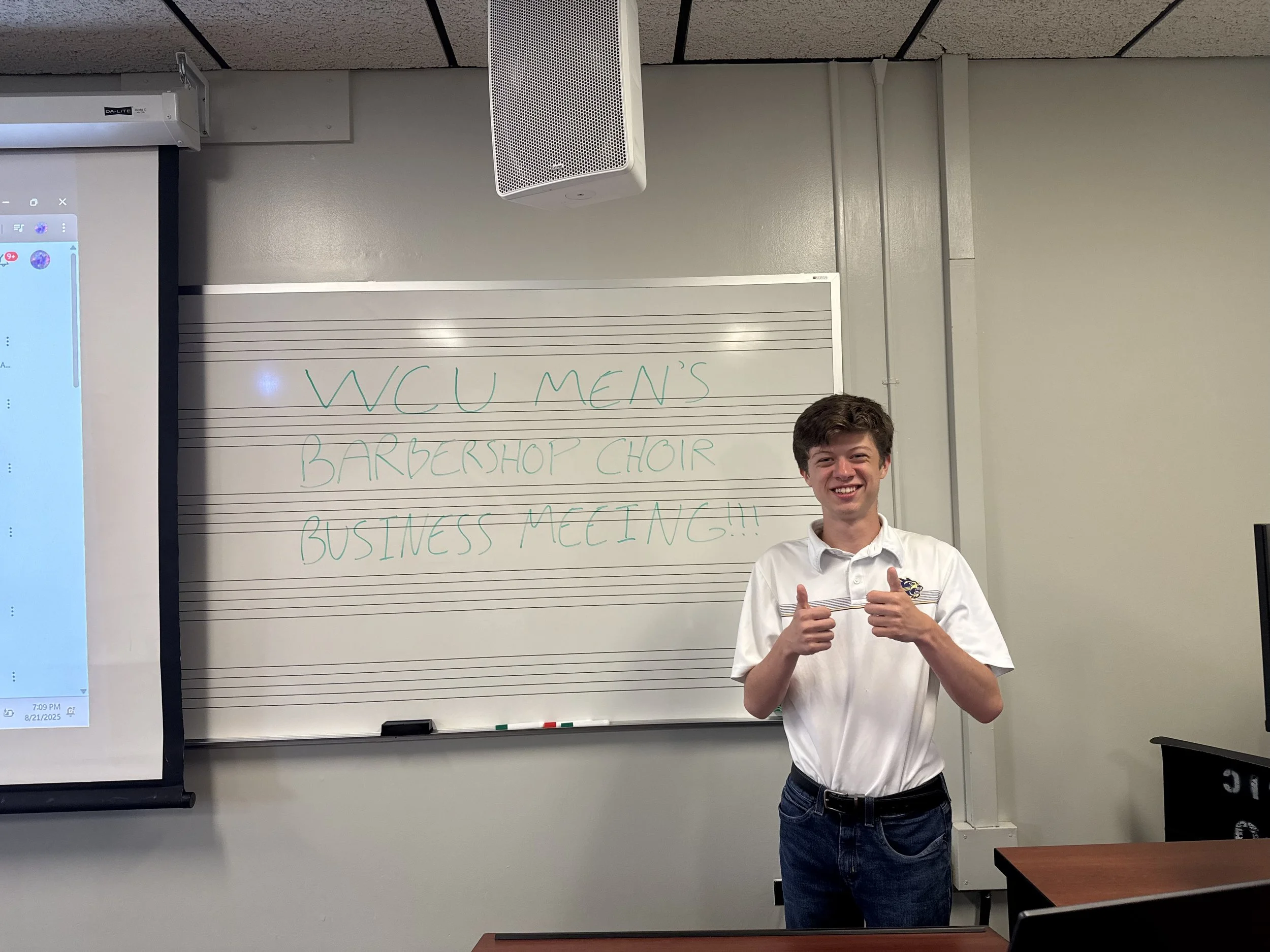 Young man standing in front of whiteboard in classroom, smiling and giving thumbs up, with message about WCW Men's Barbershop Choir Business Meeting written on whiteboard.
