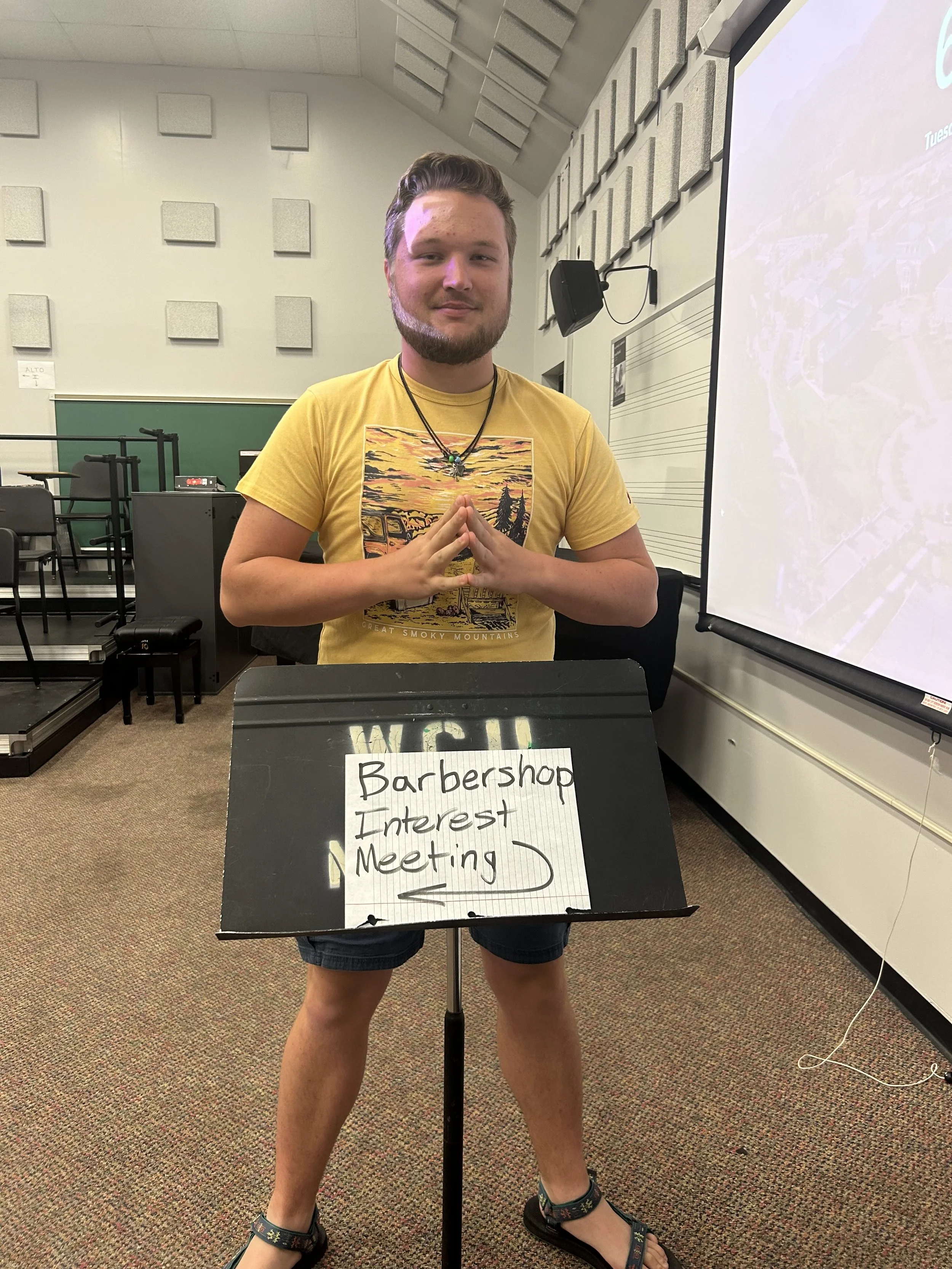A young man standing in a classroom holding a sign that says 'Barbershop Interest Meeting,' with an interest arrow pointing to the left.