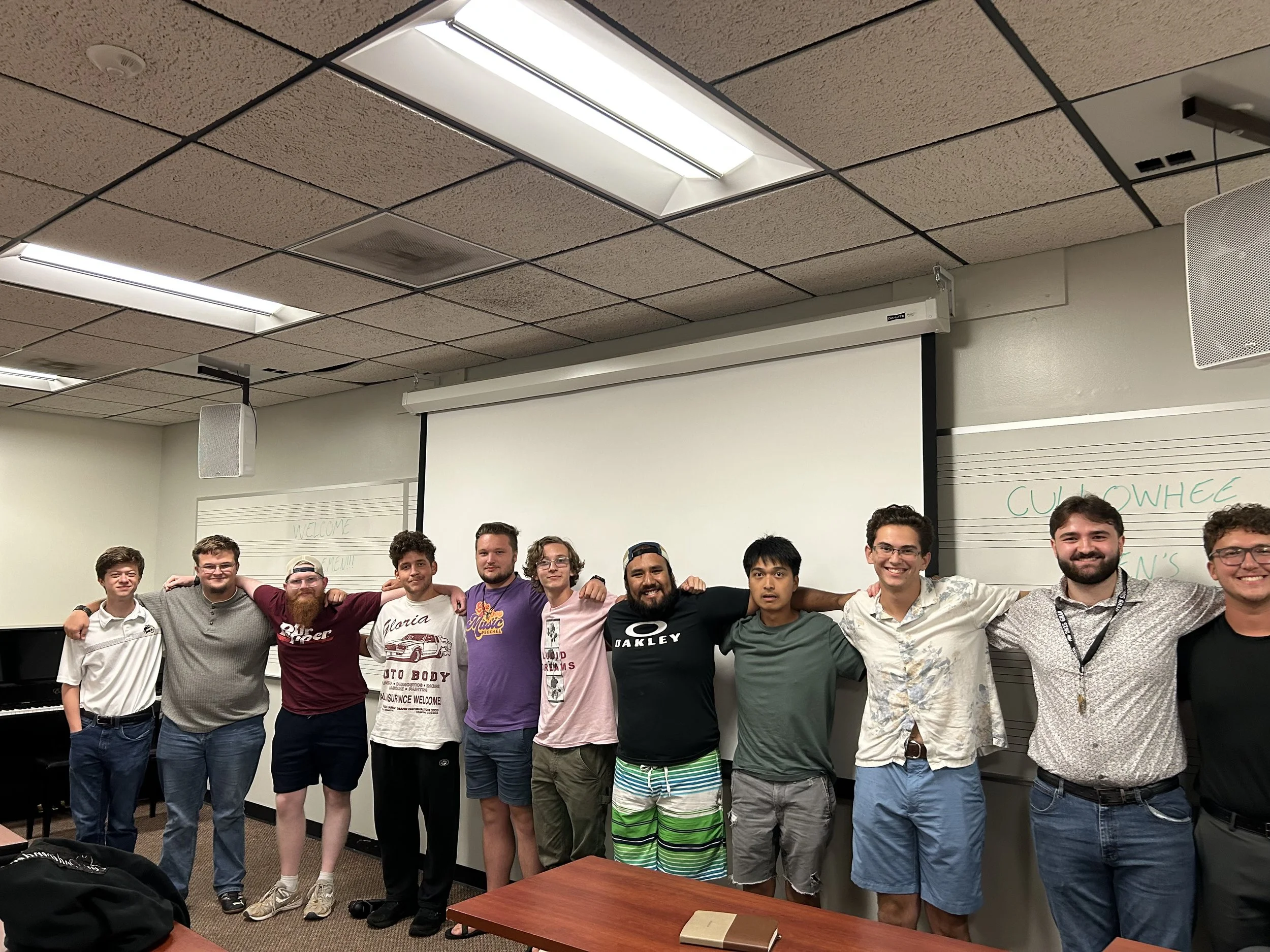 A group of eleven young men standing in a classroom with arms around each other's shoulders, smiling at the camera. The classroom has a whiteboard, a projector screen, and musical staff lines on the wall.