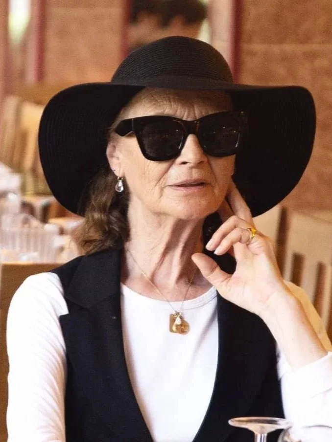 Mujer mayor con sombrero negro y ropa formal, en un ambiente interior.