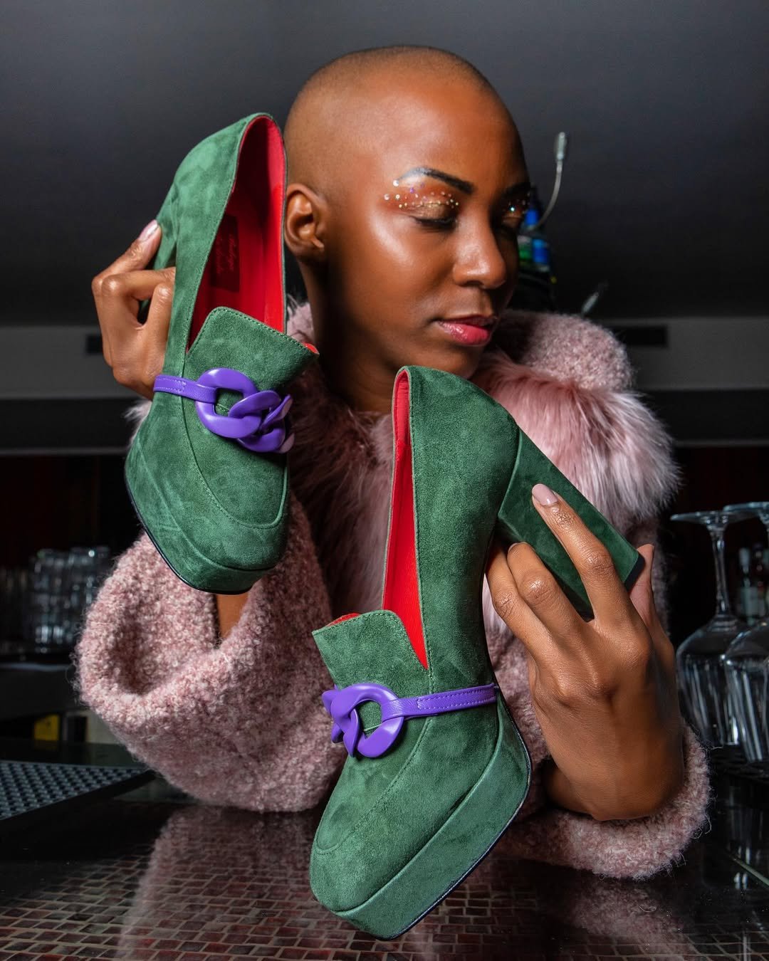 Mujer con cabeza rapada sosteniendo botas verdes con suela roja y hebillas violetas, usando abrigo rosa con forro de piel