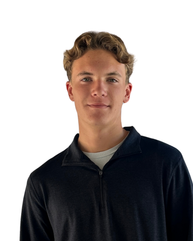 A young man with blonde hair, wearing a black zip-up jacket over a white shirt, smiling slightly against a plain white background.