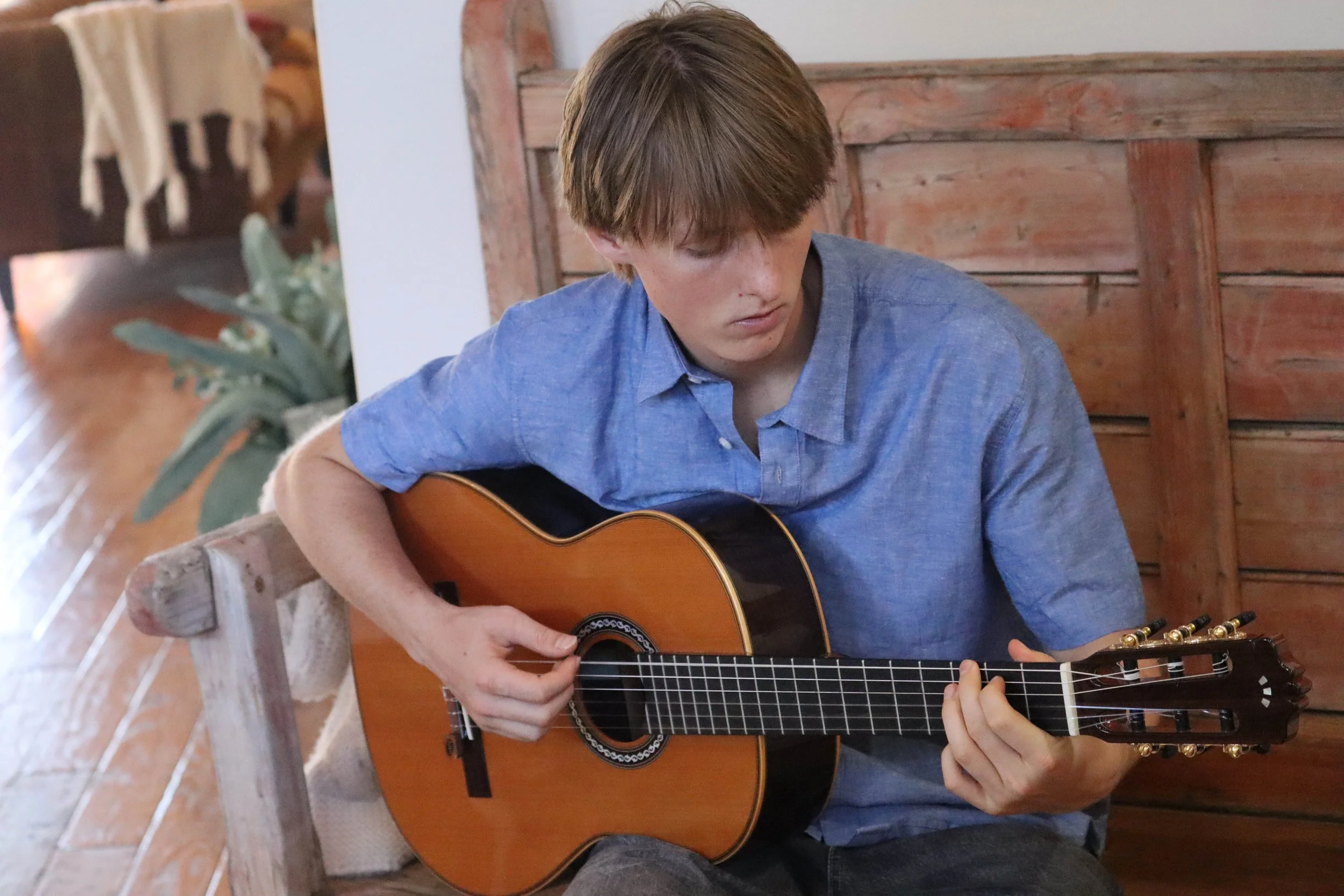 A young person with brown hair wearing a blue shirt is sitting on a wooden bench, playing an acoustic guitar.