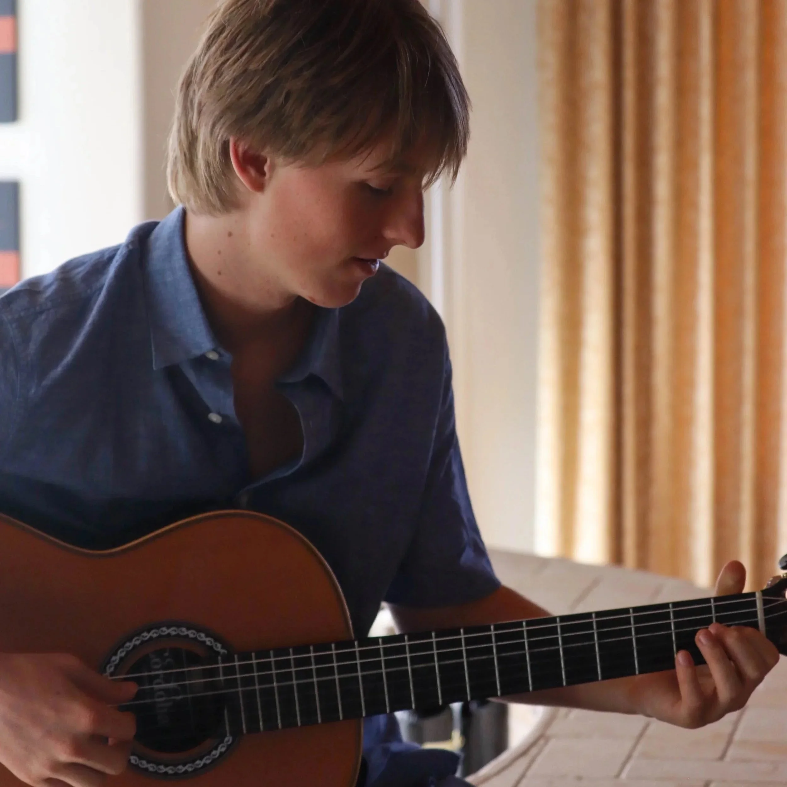 Young person with blonde hair playing an acoustic guitar in a room with beige curtains.