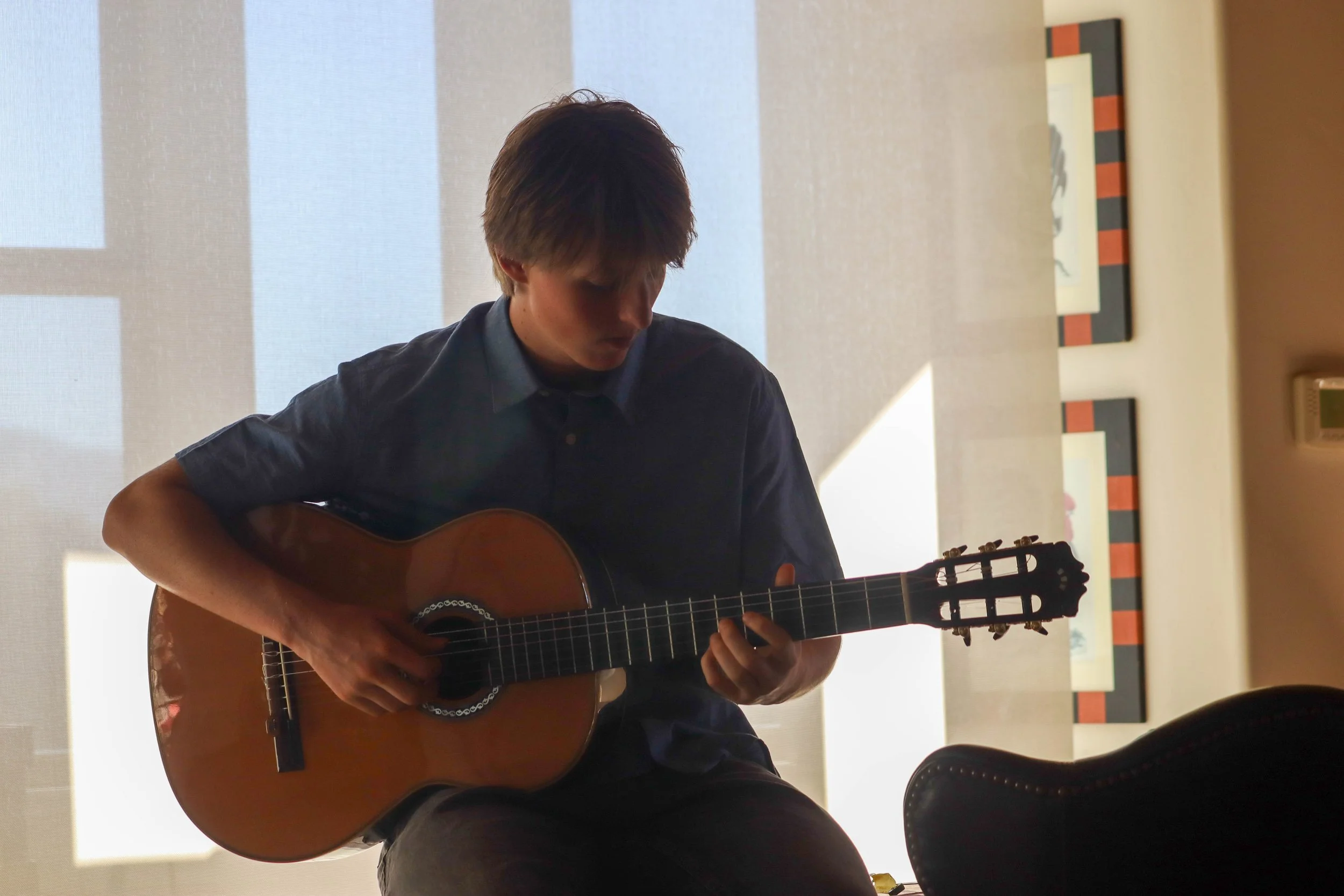 A young man playing an acoustic guitar indoors, sitting on a chair with a light-colored wall and artwork in the background.